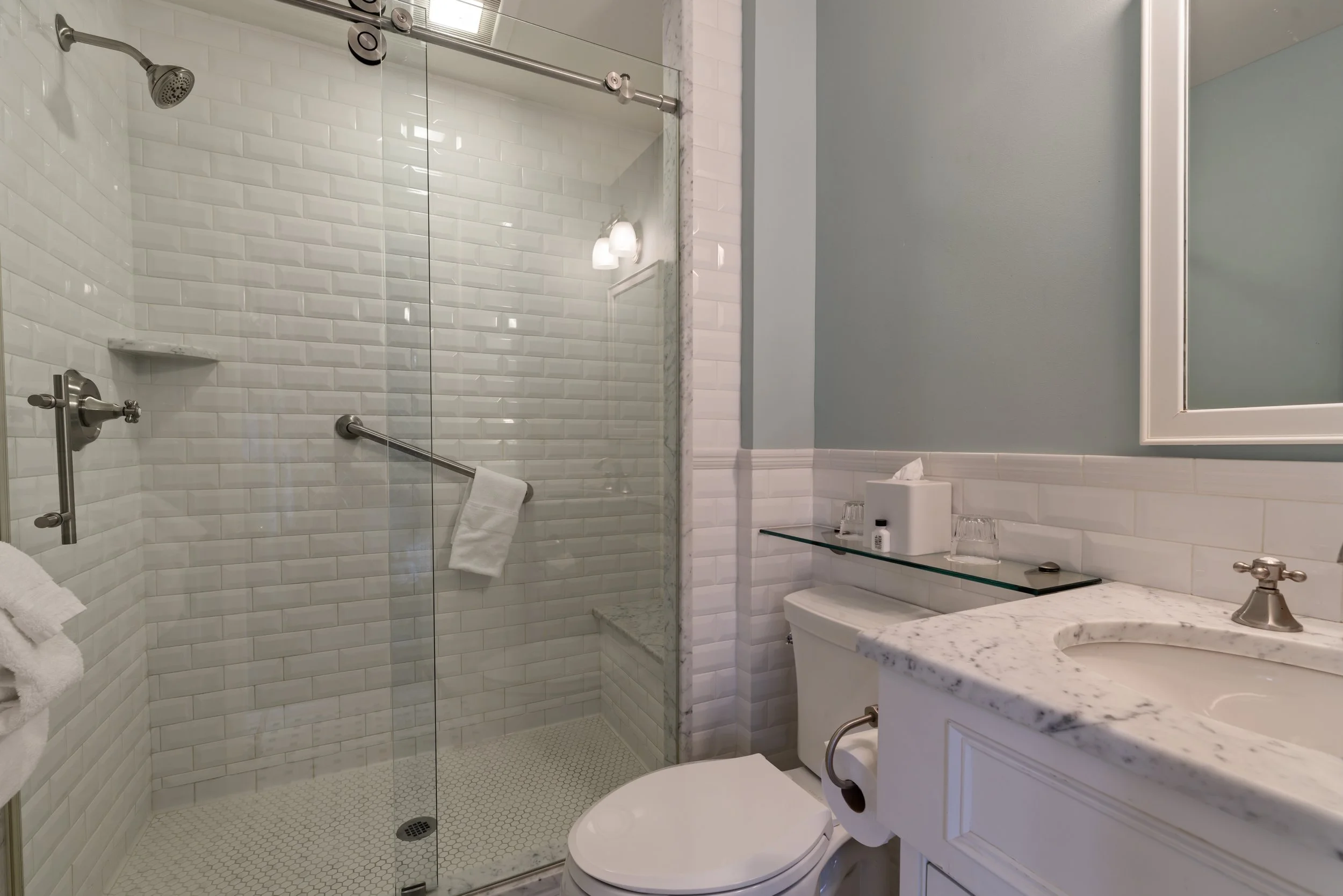 A bathroom featuring a walk-in shower with white subway tiles and a glass door, a marble countertop with a sink, a mirror, and a toilet.