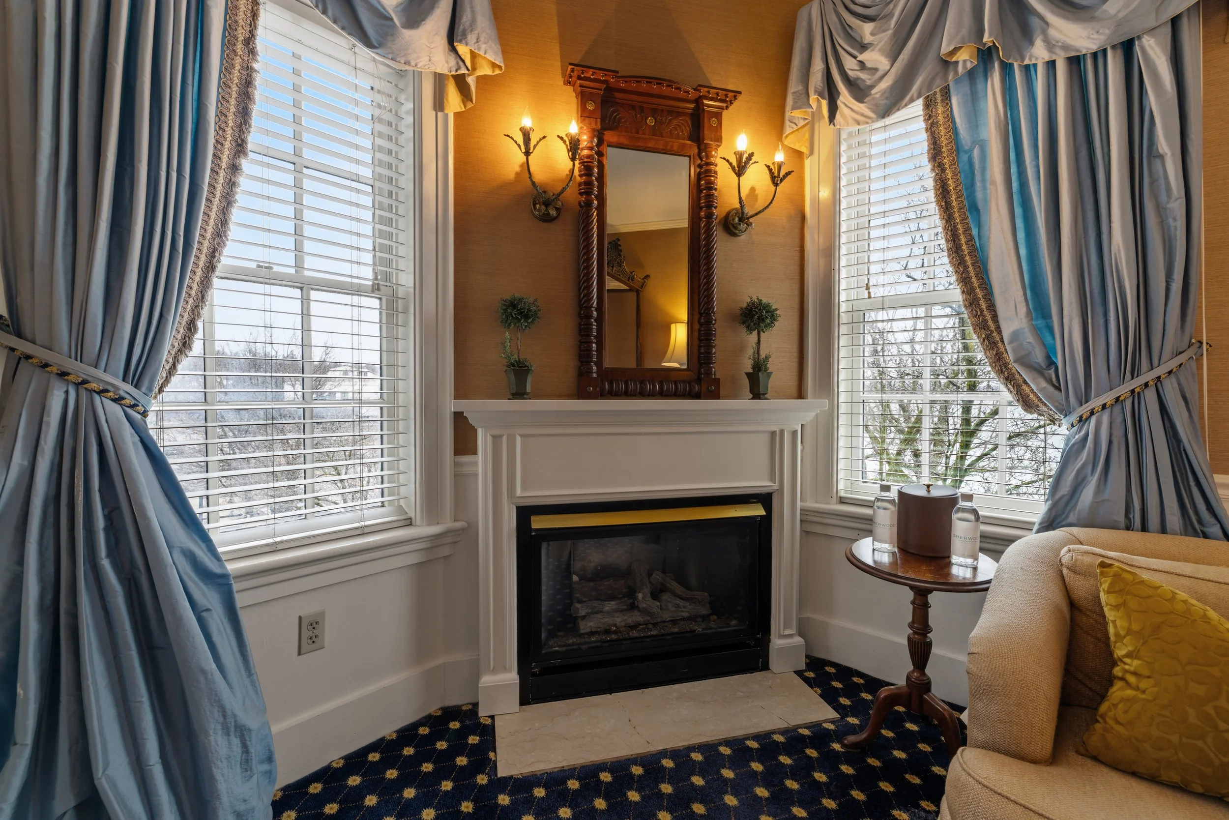 Living room with large windows dressed with blue and yellow drapes, a decorative wooden mirror above a fireplace, wall sconces, potted plants, and a beige armchair with yellow pillows.