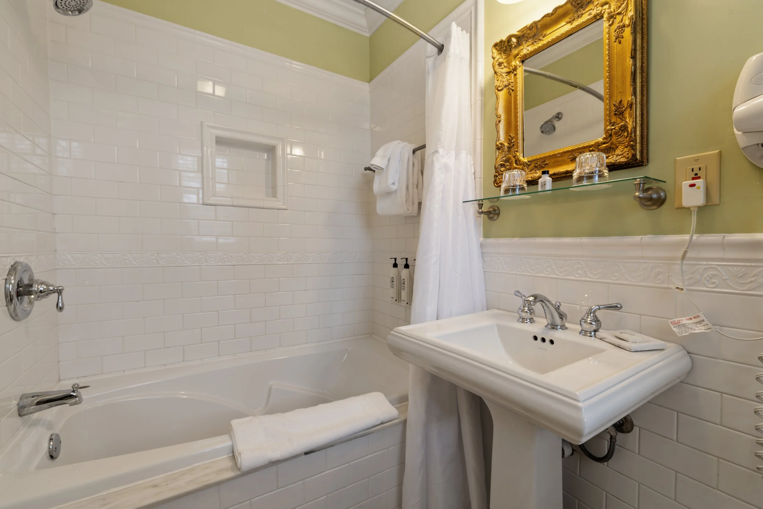 Bathroom with white tiled bathtub, shower, gold framed mirror, and green walls.