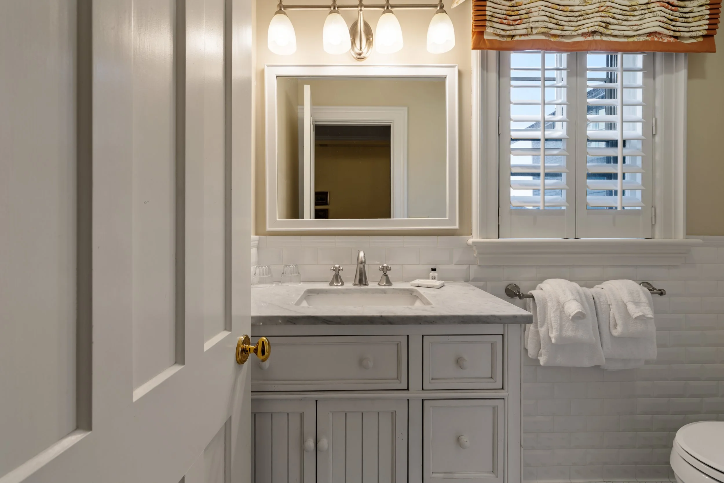 Bedroom bathroom with a white vanity, marble countertop, mirror, towel rack, and window with blinds.