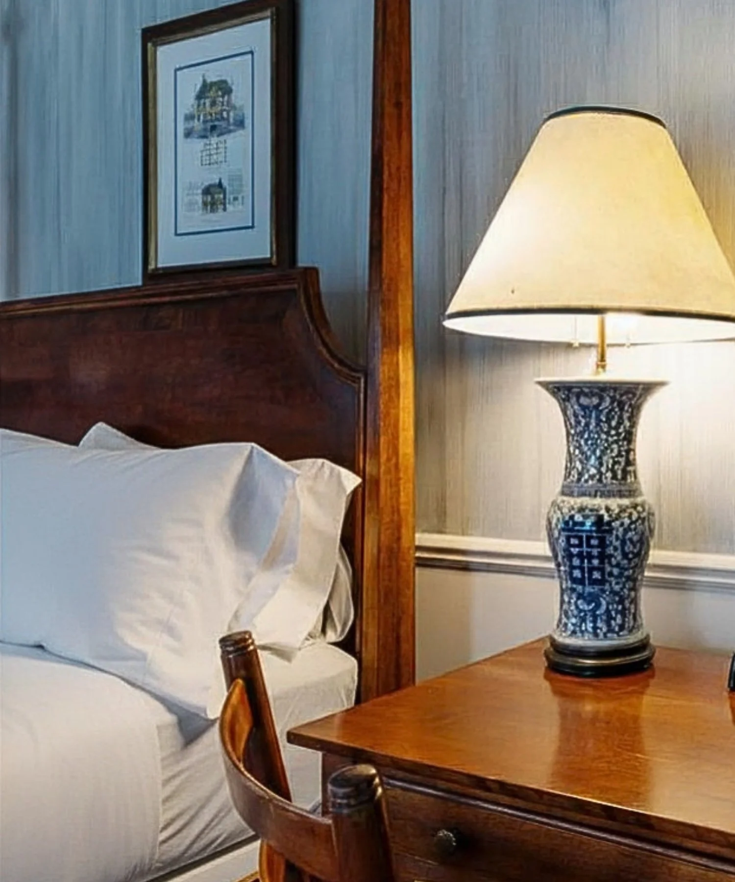 Interior of a room with a wooden bedpost, a bed with white sheets and pillows, a wooden dresser, a framed picture on the wall, a decorative blue and white porcelain lamp on a wooden side table, and chairs nearby.