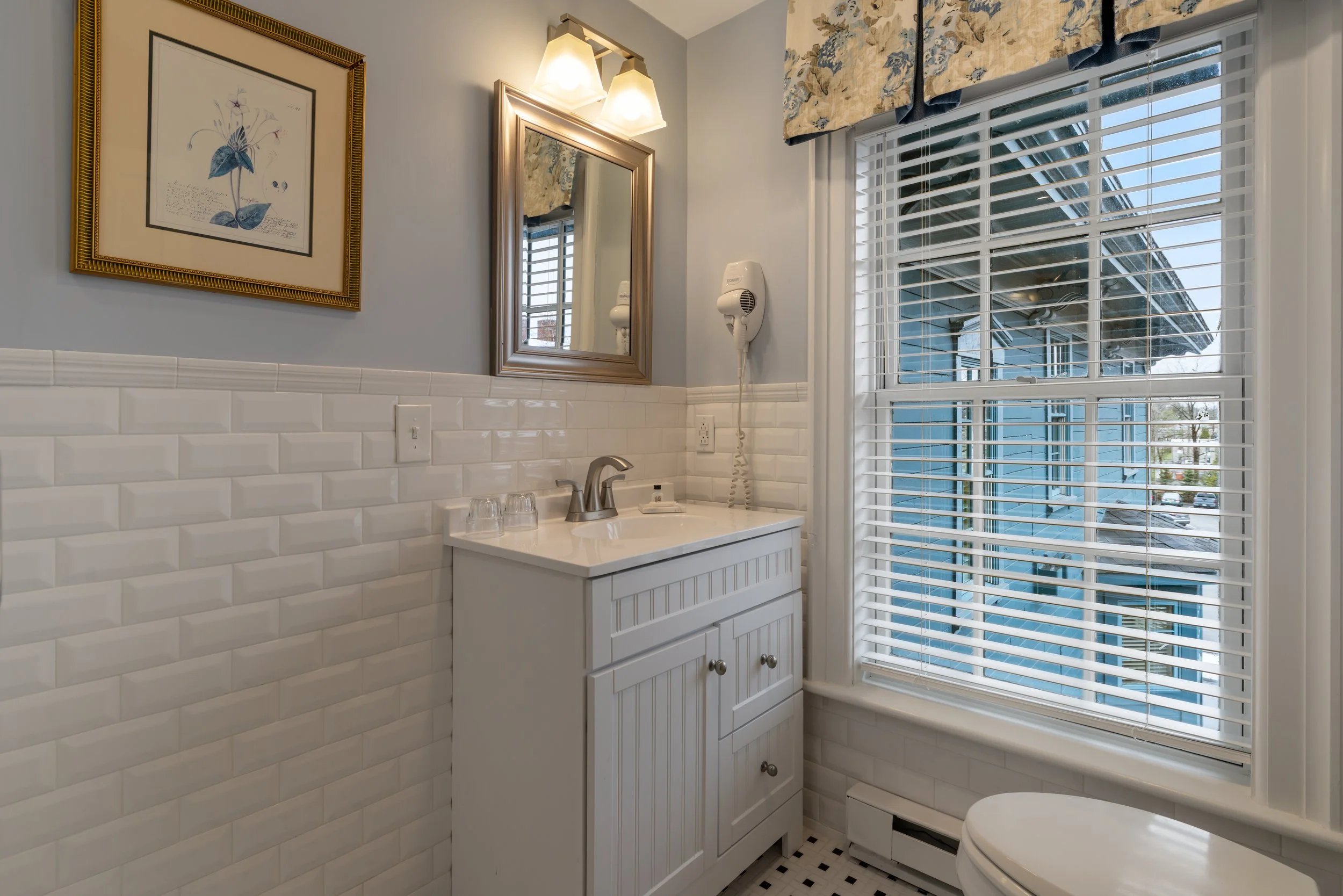 Bathroom with white subway tile wainscoting, small black and white tiled floor, white vanity with a sink, large window with blinds and floral valance, mirror, wall-mounted hairdryer, artwork, and wall-mounted light fixture.
