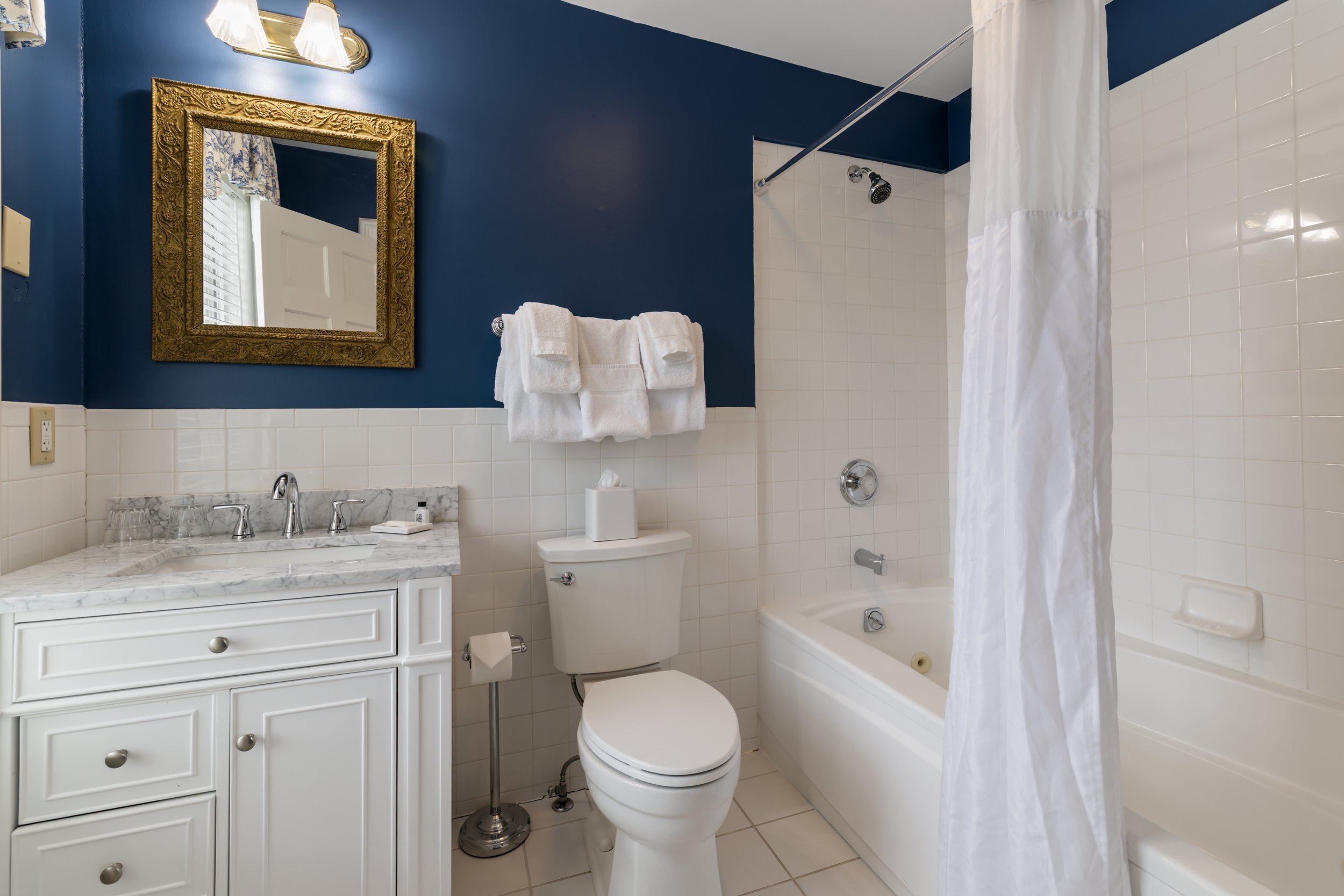 A bathroom with blue walls, white tile wainscoting, a white vanity with a marble countertop, a mirror with an ornate gold frame, a wall-mounted light fixture, a white towel on a towel rack, a toilet, and a bathtub with a curved shower curtain rod and white shower curtain.