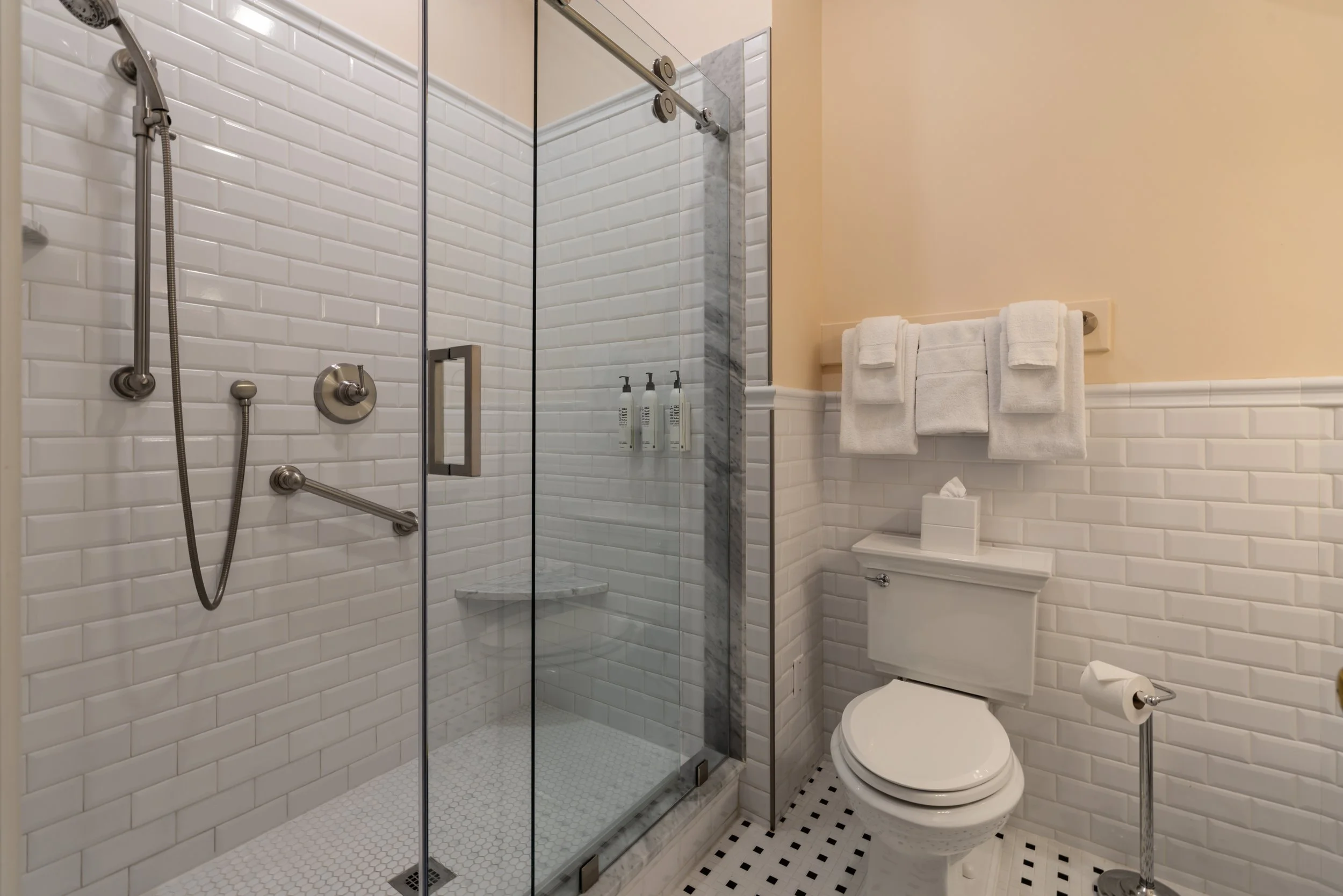 A bathroom with a glass shower enclosure, white subway tile walls, and a white toilet. There are black and white checkered floor tiles, and a towel rack with several white towels. Inside the shower, there are bottles of toiletries on a built-in shelf.