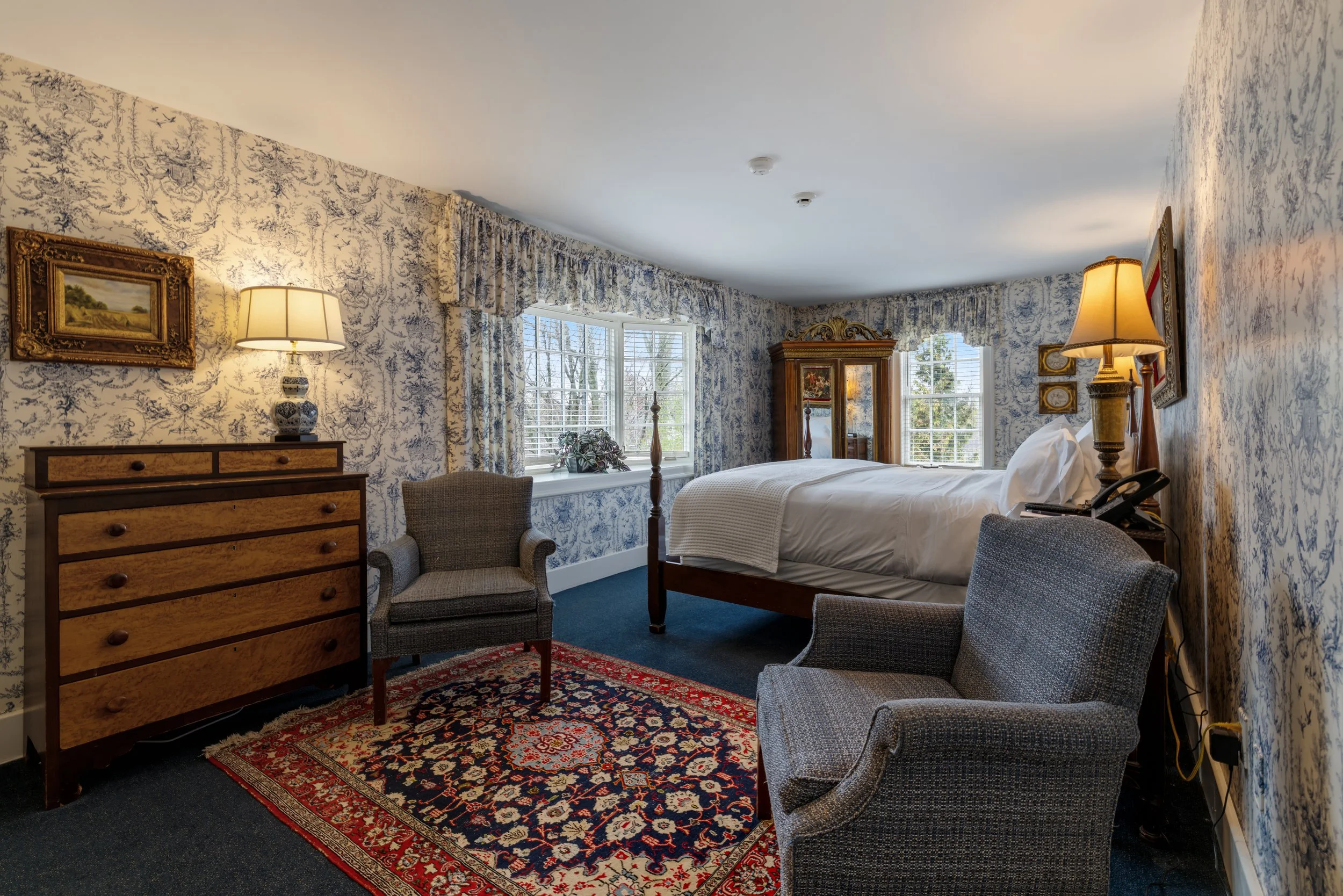 A cozy bedroom with patterned wallpaper, a large bed, two lamps, a wooden dresser, and several chairs. Large windows with blue and white patterned curtains let in natural light, and a bookcase or armoire is visible in the corner.
