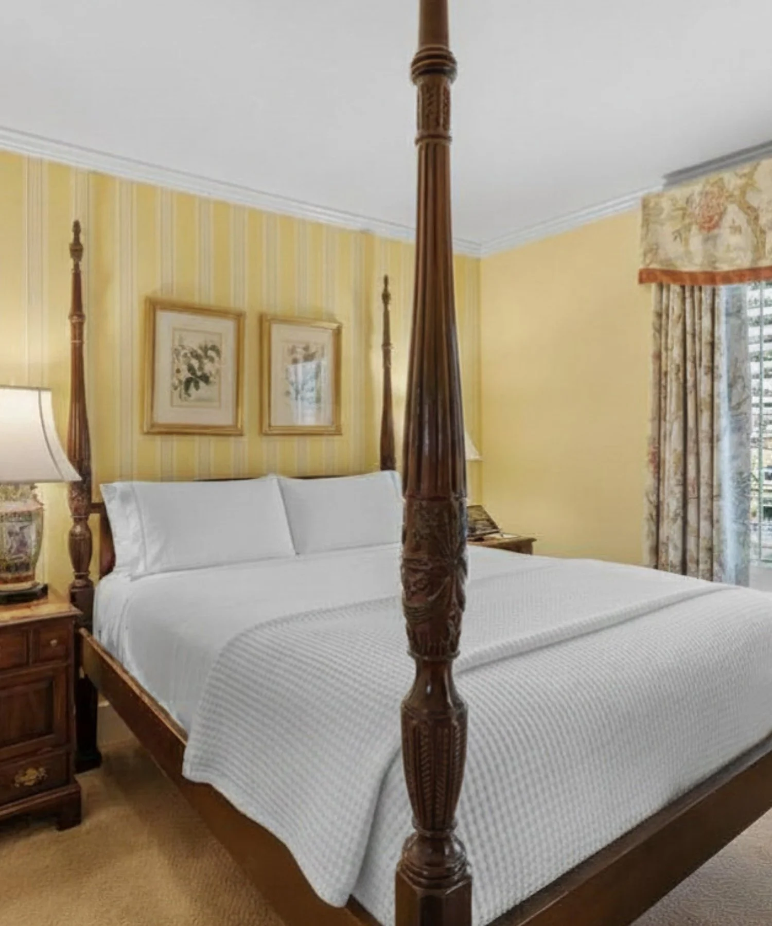 A bedroom with a four-poster bed, white bedding, a wooden nightstand, a lamp, framed art on yellow striped walls, and window curtains with a floral pattern.