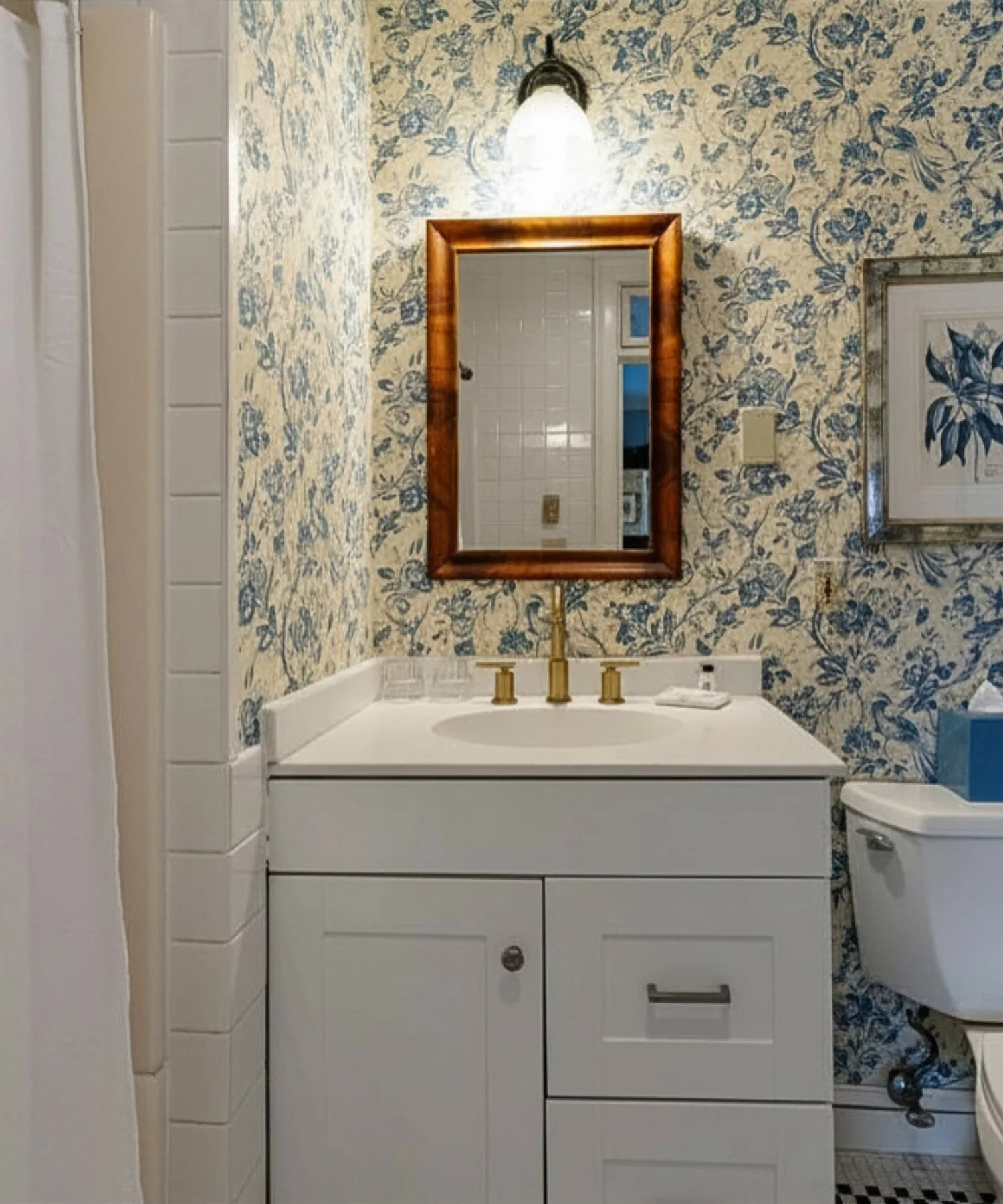 A bathroom vanity with a white cabinet and drawers, gold faucet, and mirror above. Wallpaper with blue floral pattern on beige background, wall-mounted light fixture, and a framed picture on the wall. Toilet nearby with a box of tissues on top.