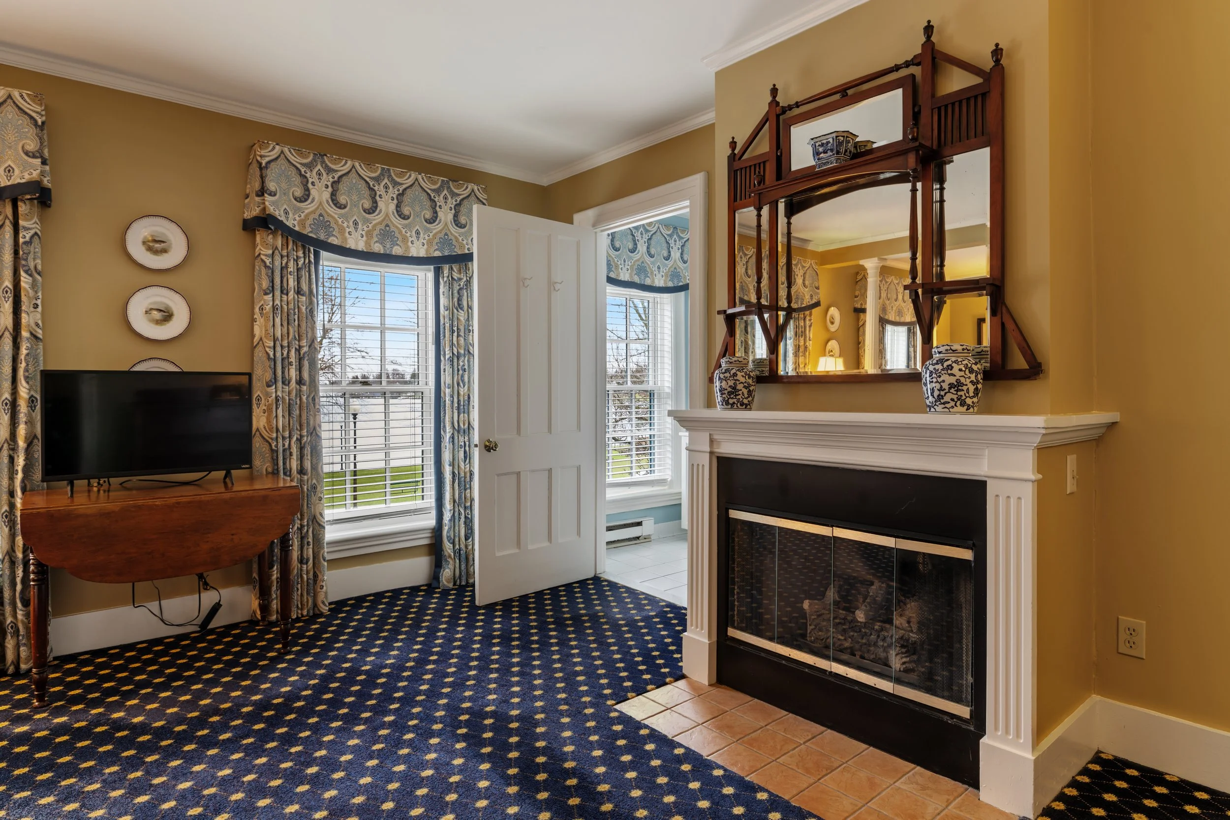 Interior of a living room with two large windows, patterned curtains, a wall-mounted mirror, a fireplace with decorative vases on the mantel, a small wooden table with a TV, and a patterned carpet.