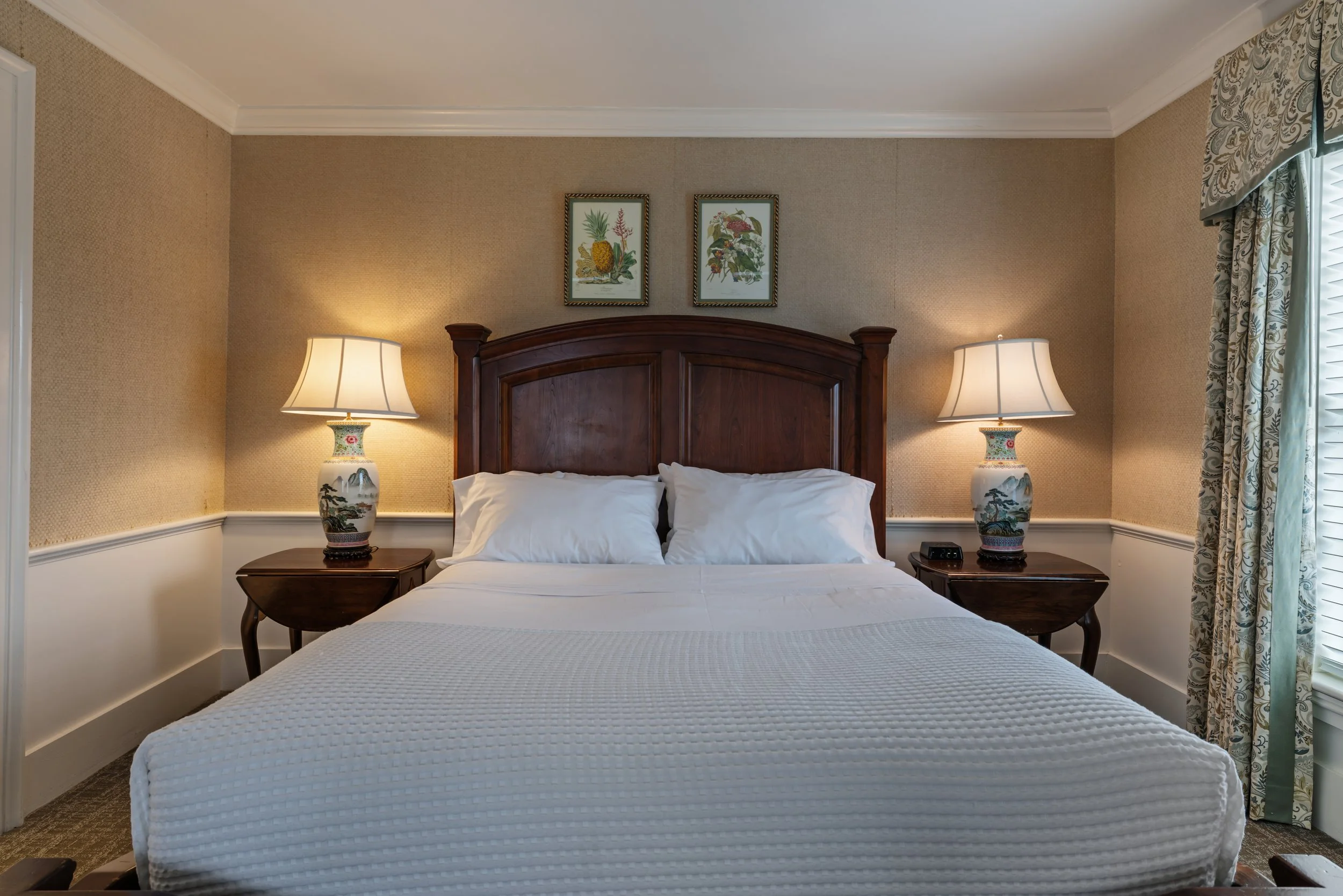 A bedroom with a wooden headboard, white bedding, and two decorative table lamps on bedside tables. There are framed botanical illustrations on the wall above the headboard and patterned curtains on the window to the right.