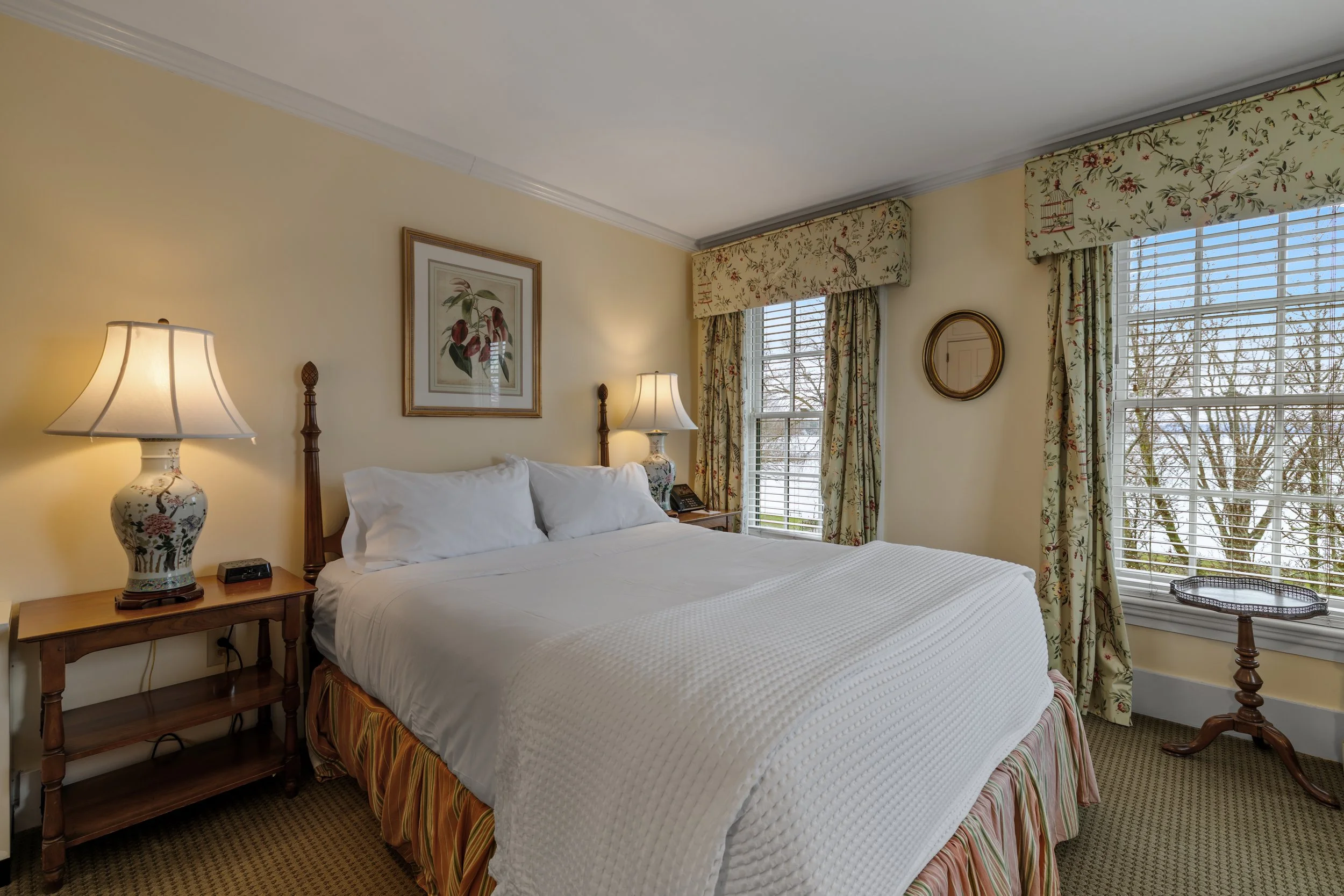 A cozy bedroom featuring a large bed with white bedding and a striped bed skirt, accompanied by wooden bedside tables with floral lamp bases and lampshades, floral curtains and valances, a picture frame of botanical art on the wall, and a small round mirror, near large windows with blinds showing trees outside.