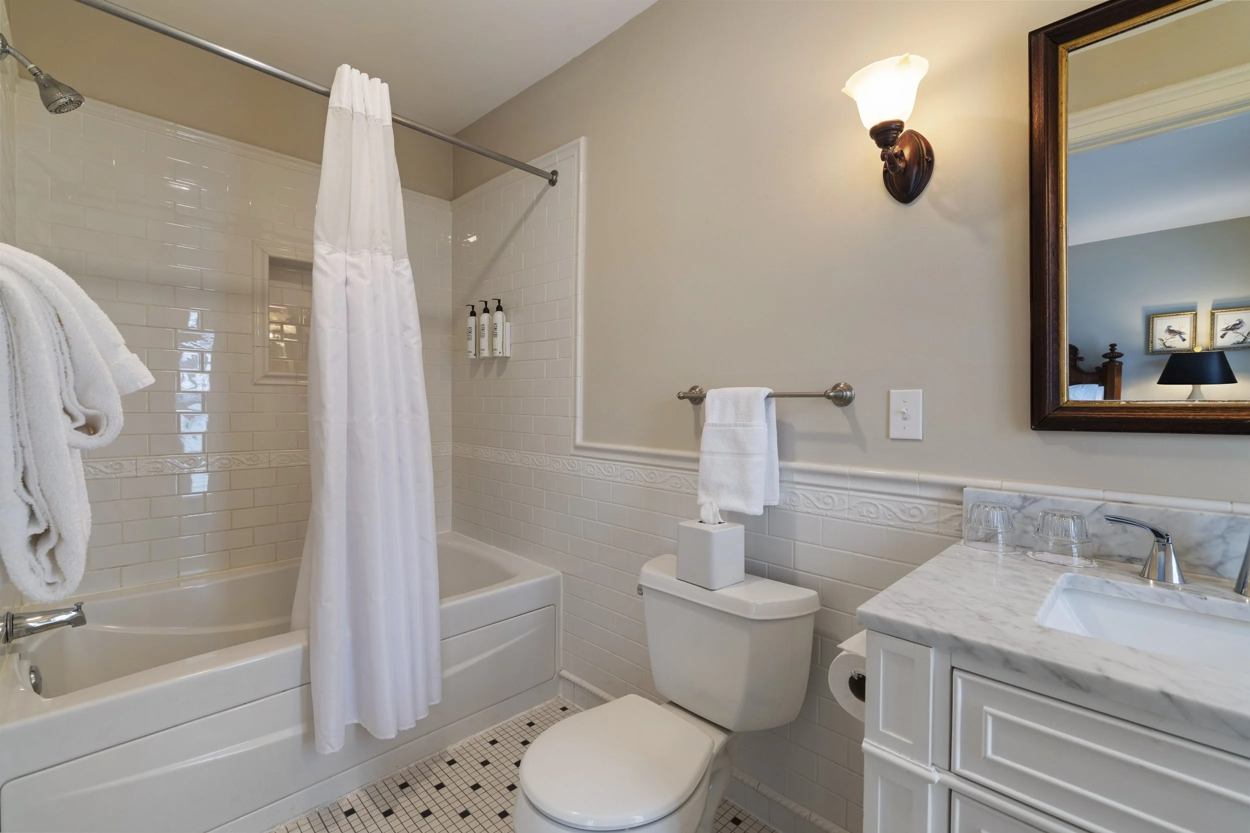 A bathroom with a bathtub and shower, white tiled walls, a toilet, a marble counter with a glass, a mirror, a wall-mounted lamp, and a towel rail with towels.
