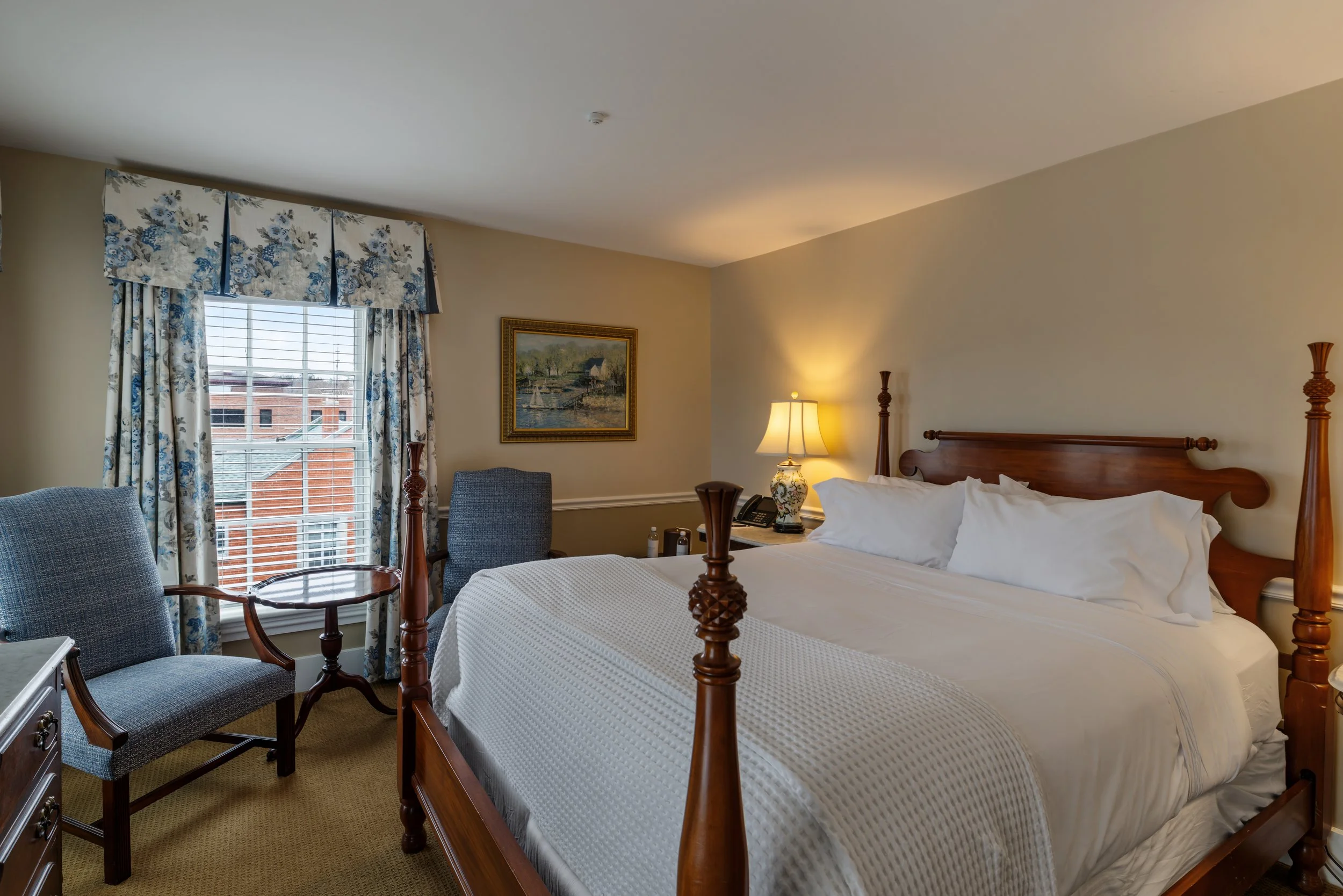 A hotel room with a large bed, wooden bed frame, nightstand with a lamp, two chairs, a window with floral curtains, and a framed painting on the wall.