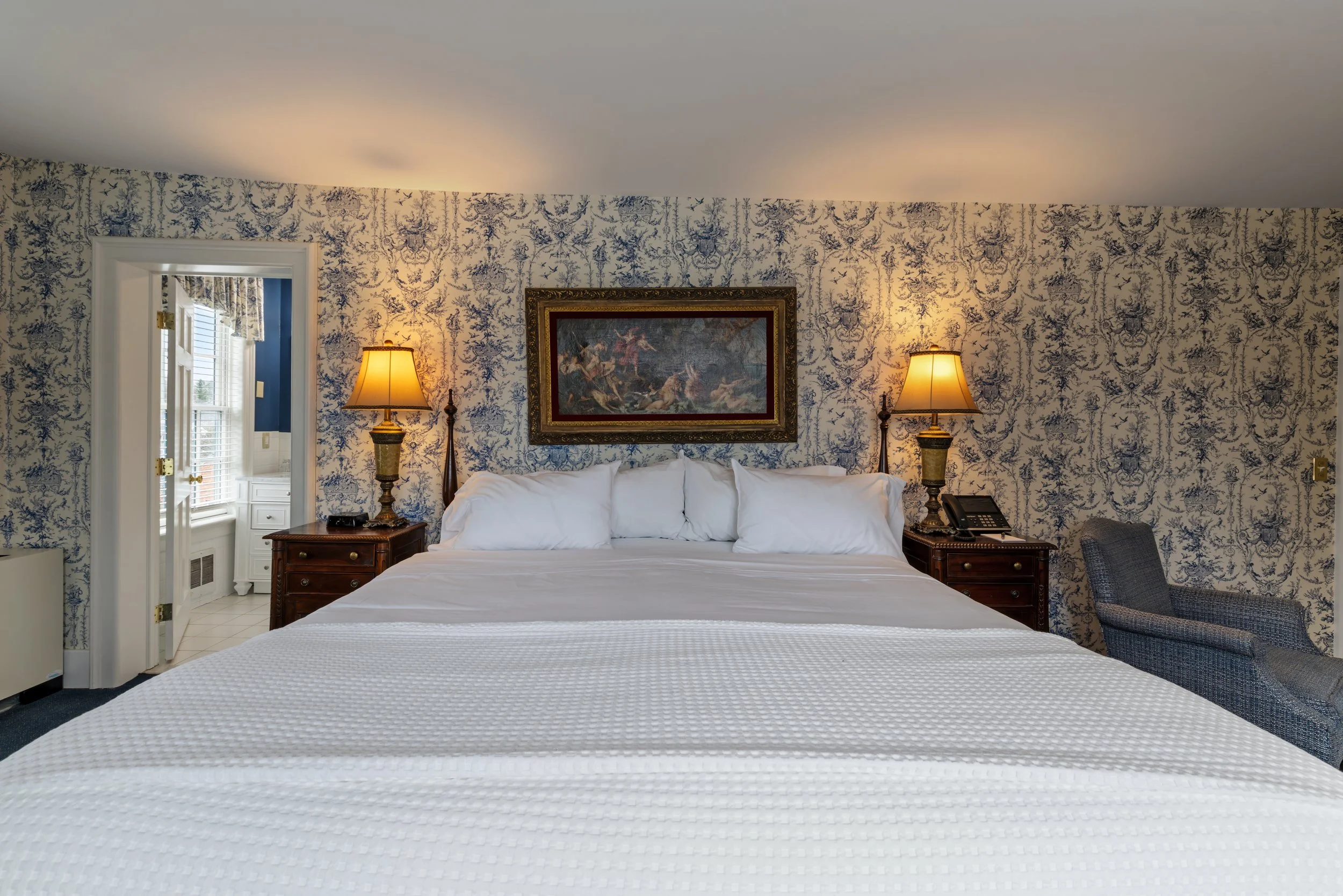 A hotel room with a bed, two bedside lamps, a framed painting, and a patterned wallpaper background.