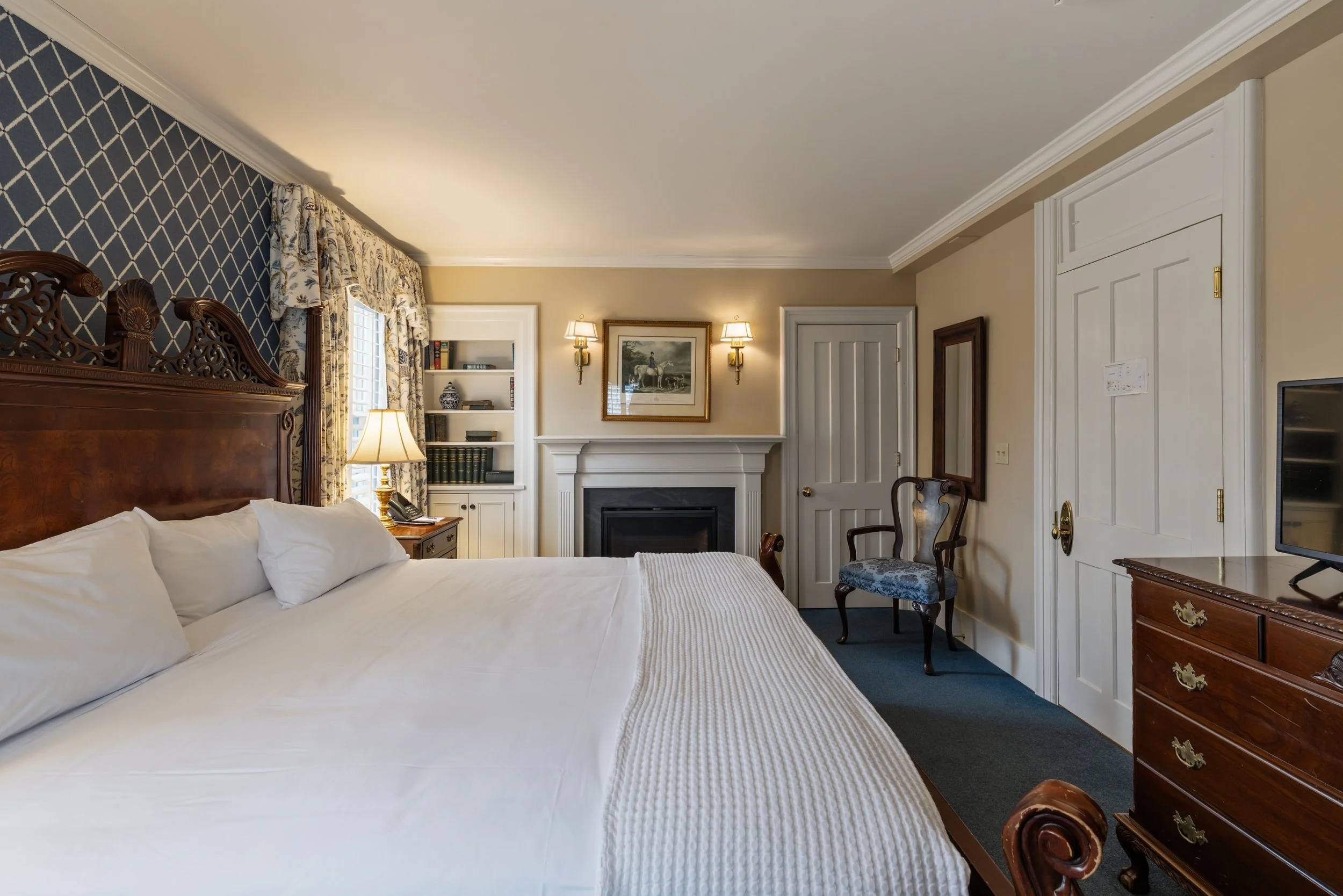 Interior of a traditional bedroom with a wooden headboard, white bedding, a bedside lamp, a bookshelf, a fireplace, framed artwork, wall sconces, a mirror, and a dresser.
