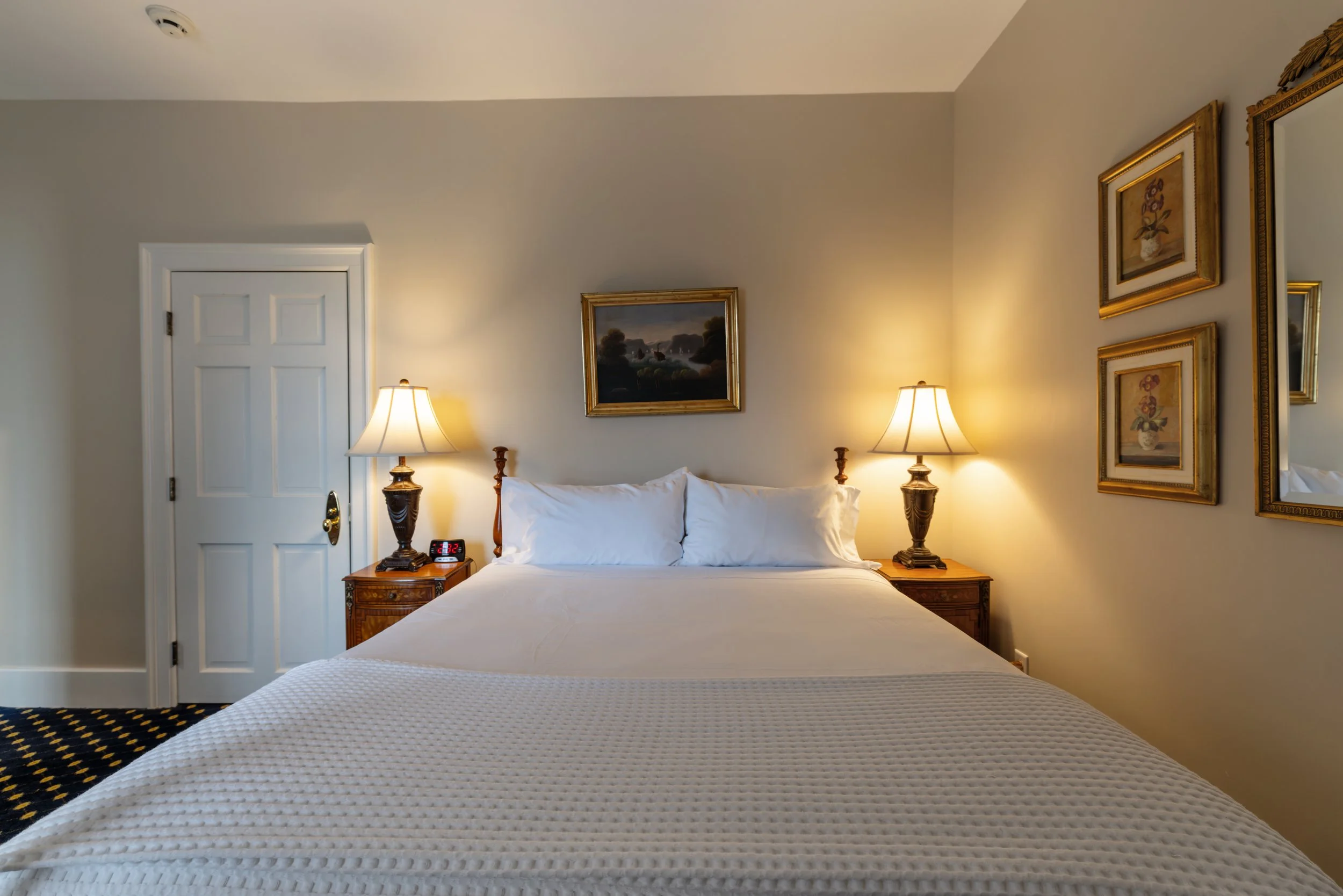 A hotel room featuring a bed with white bedding, wooden nightstands with lamps on either side, a painting above the bed, and framed artwork on the adjacent wall.