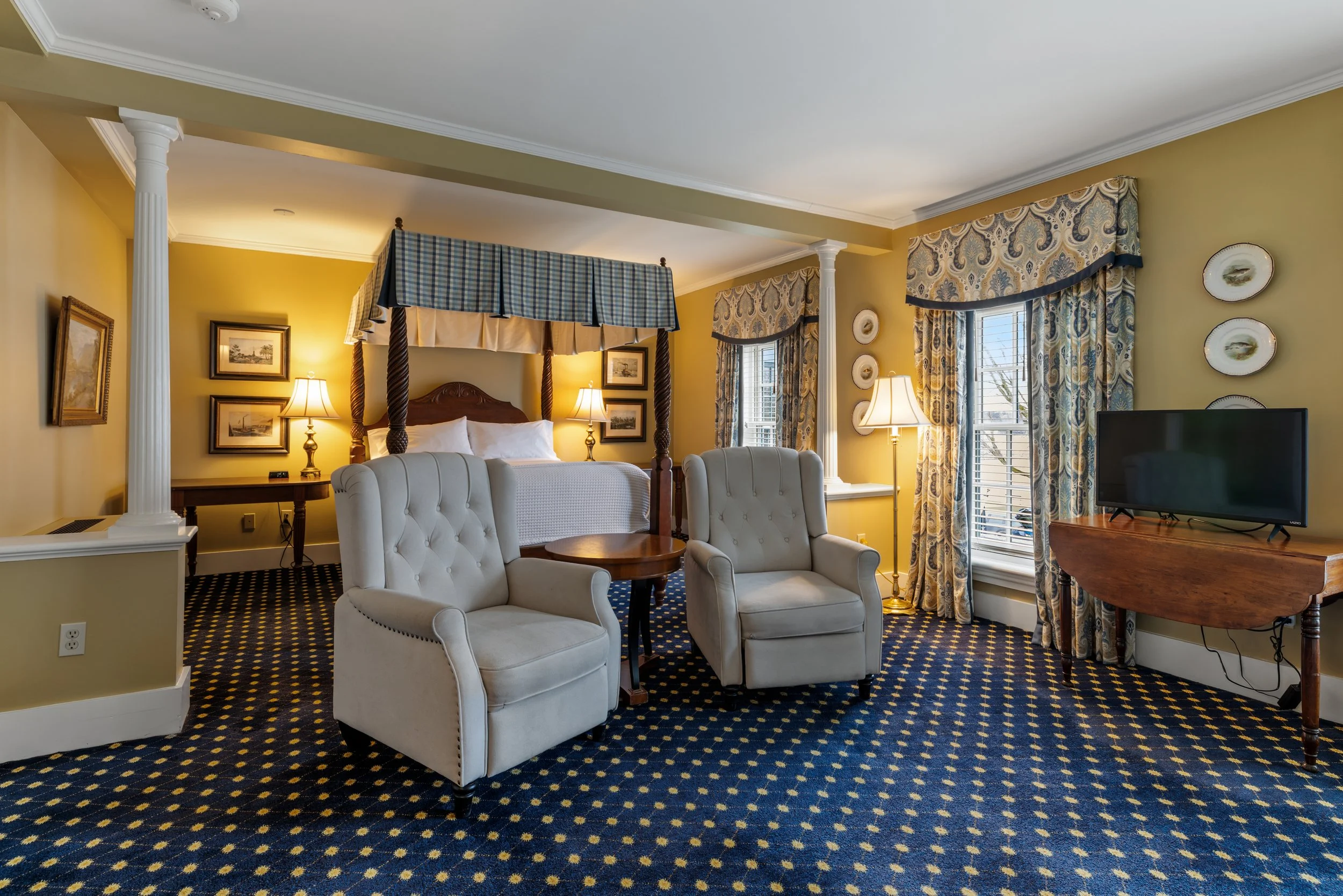 Interior of a traditional hotel room with white upholstered armchairs, a four-poster bed with wooden posts and blue checkered canopy, yellow walls adorned with framed artwork and decorative plates, gold and cream lamps, windows with patterned curtains, and a wooden table with a television.