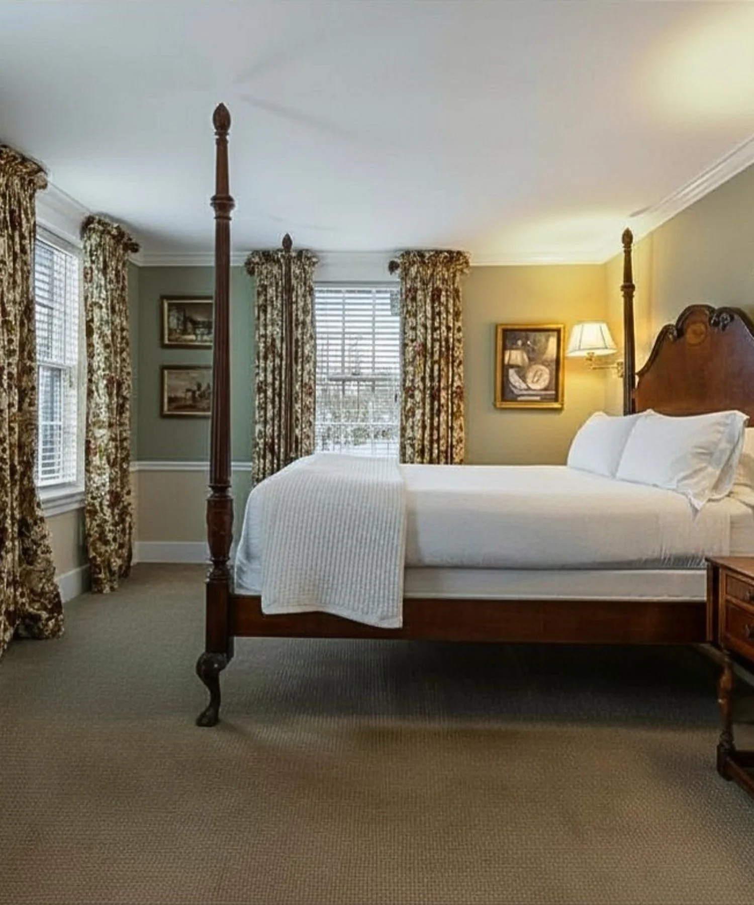 A bedroom with a four-poster bed with white bedding, floral patterned curtains, two windows, framed pictures on the wall, and a lamp.