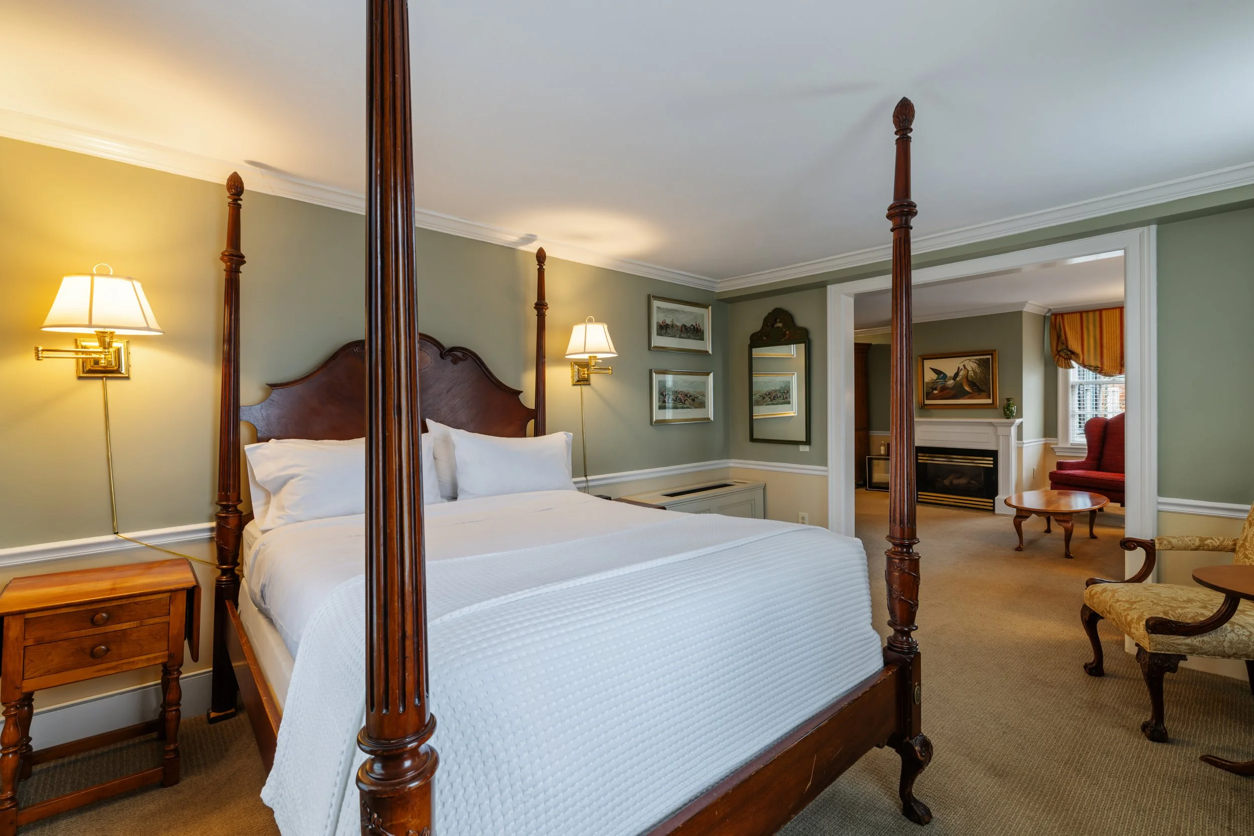 A bedroom with a four-poster bed with white bedding and pillows, wooden nightstands, and wall-mounted lamps. The room has framed artwork on the walls, a mirror, a fireplace, and a window with valance curtains. There is an armchair with an embroidered cushion and a small round wooden side table.