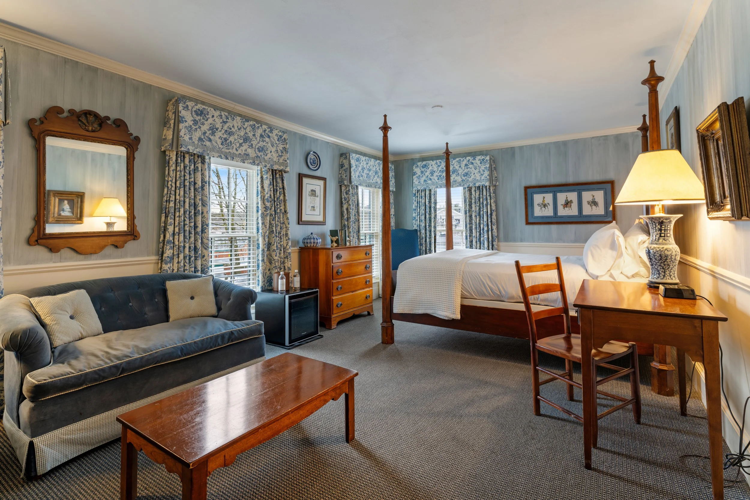 A cozy bedroom with a four-poster bed, blue wallpaper, a wooden dresser, framed artwork, a table lamp, a blue armchair, and a sofa with pillows. Multiple windows with floral curtains allow natural light.