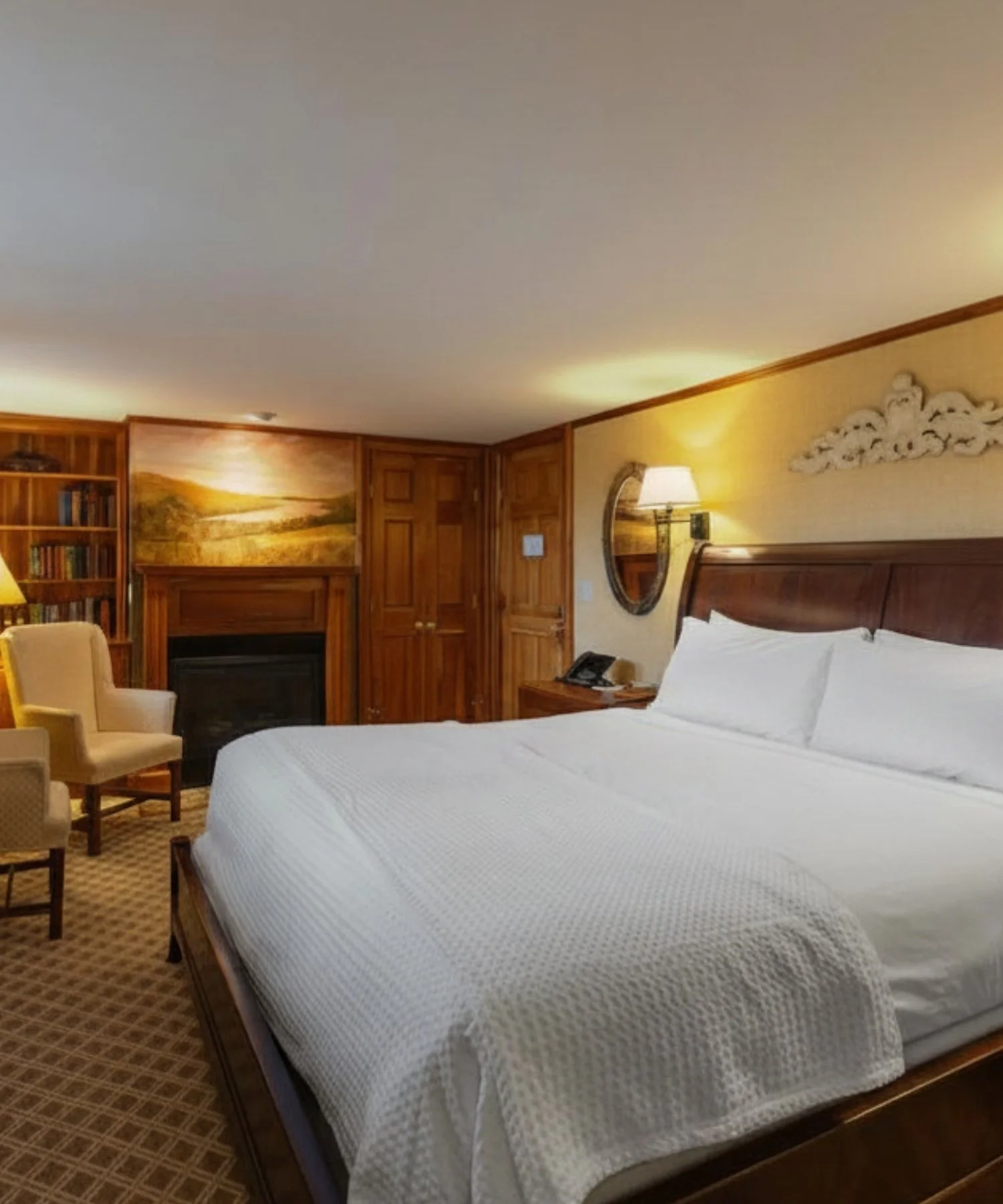 A hotel room with two beds, a wooden nightstand, a table lamp, a decorative wall clock, an armchair, a bookshelf with books, a landscape painting, and a fireplace.