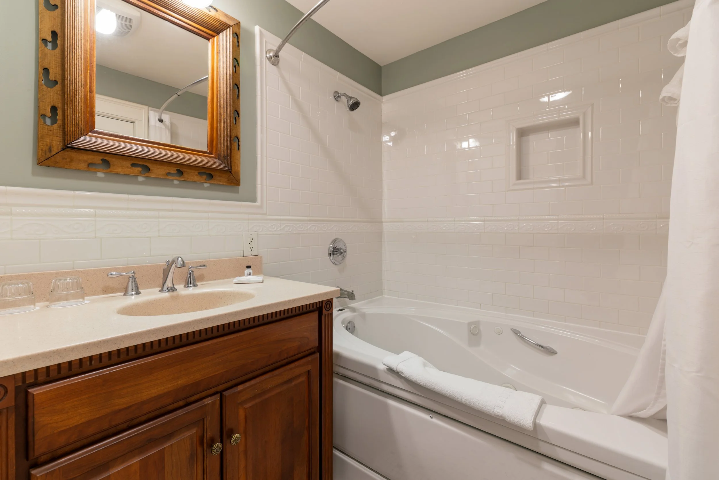Bathroom with a wooden-framed mirror above the sink, a countertop with a faucet, and glasses. Next to the sink is a bathtub with a white towel draped over its edge, a towel bar, and a small recessed shelf in white tiled wall. The wall behind the bathtub is tiled in white with a decorative border.