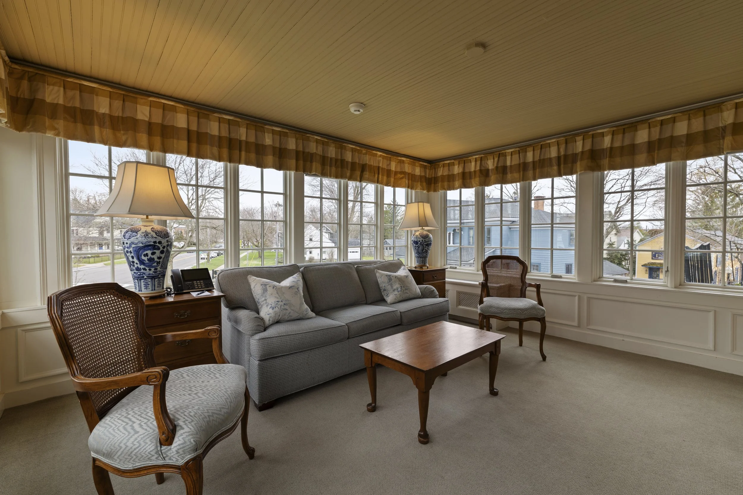 Living room with large windows, two table lamps with blue and white ceramic bases, a gray sofa, and wooden chairs.