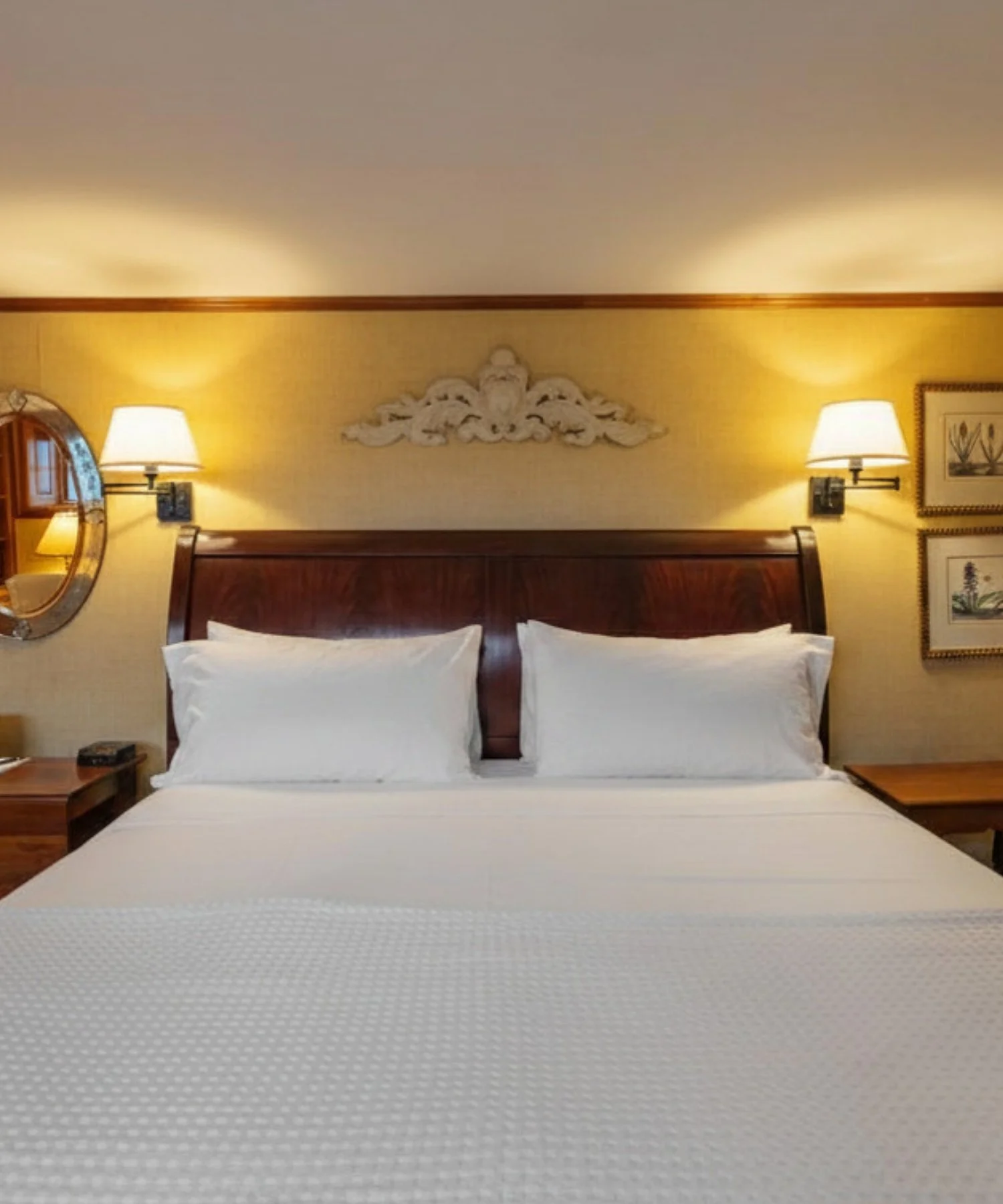 Hotel room with neatly made bed, wooden headboard, two white pillows, wall-mounted lamps on each side, decorative wall art, and a mirror on the left side.