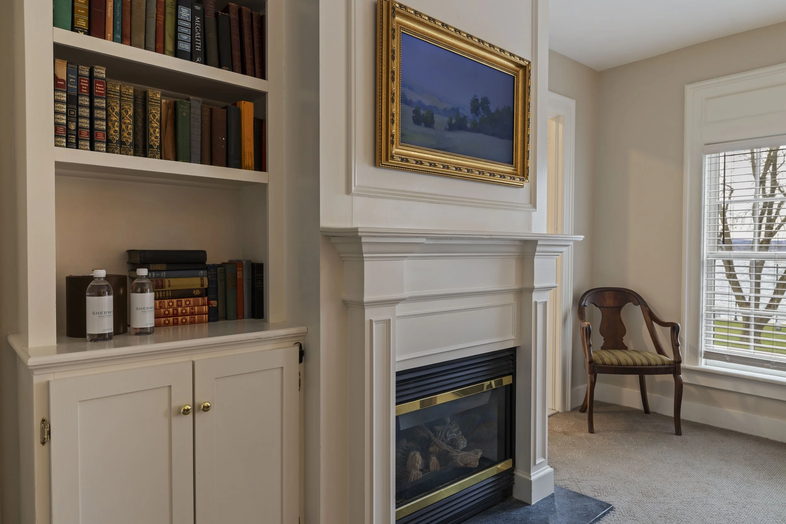 A corner of a room featuring a white built-in bookshelf with multiple shelves holding books and two bottled water. There is a white fireplace with a decorative gold frame mirror hung above it. To the right, there is a window with white blinds showing trees outside and a wooden chair with a striped cushion near the window.