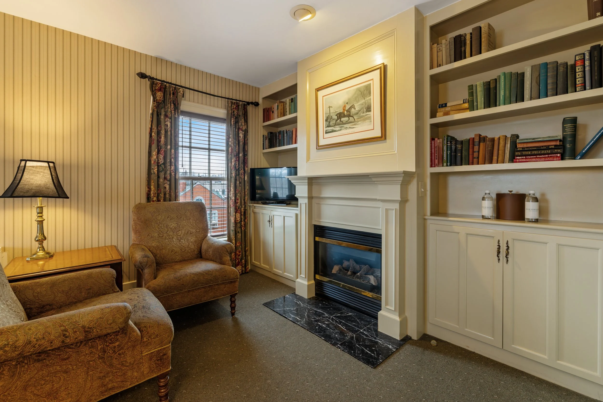 Living room with beige patterned armchairs, a small wooden table, a lamp, floral curtains, a window with blinds, bookshelves, a framed picture of a person riding a horse, a fireplace with a black marble hearth, and decorative bottles.