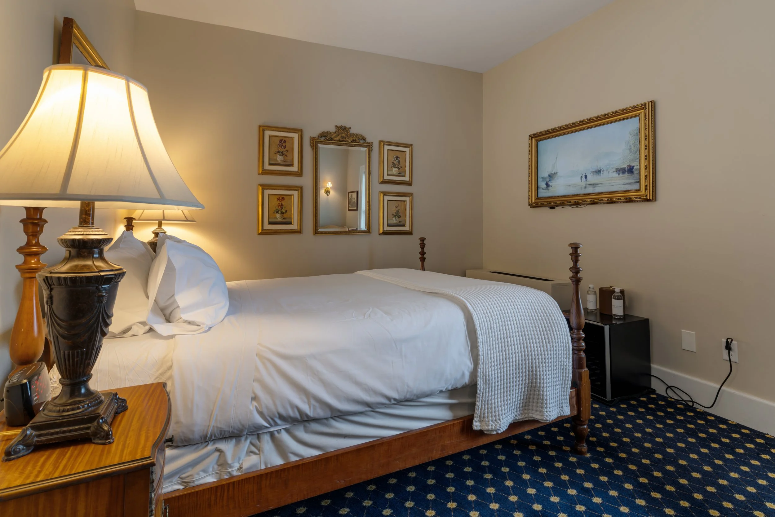 A hotel room with a wooden bed frame, white bedding, bedside table with a traditional lamp, framed artwork on cream walls, a mirror, and a small black refrigerator with water bottles, with a blue patterned carpet.