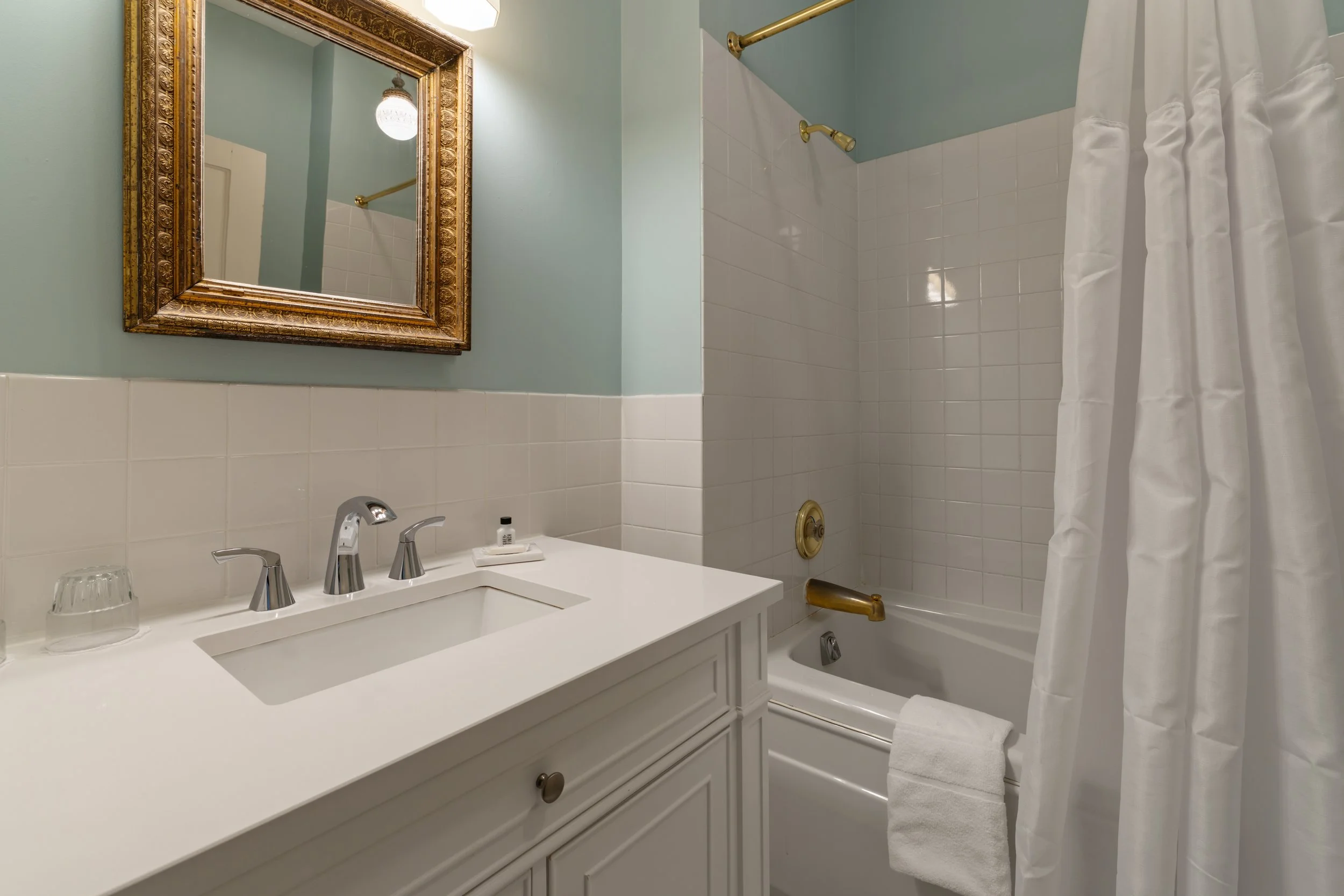 A bathroom with a white vanity featuring two faucets and two glasses. A large ornate gold-framed mirror hangs above the sink. Behind the vanity, light blue walls and white tiled wall are visible. To the right, a bathtub with a white shower curtain and gold fixtures, including a showerhead and faucet, are visible. A white towel hangs over the tub's edge.