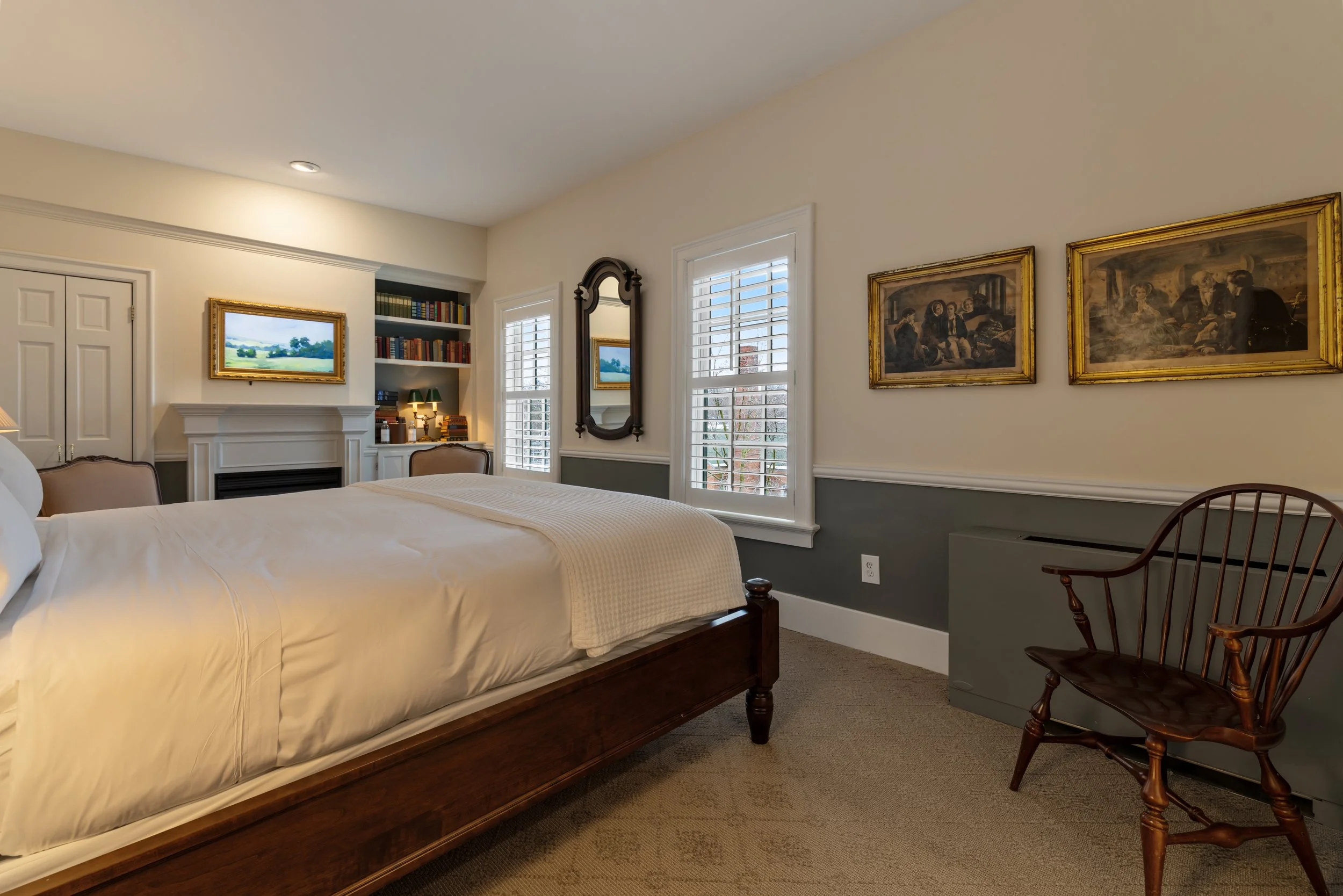A cozy bedroom with a wooden bed frame, white bedding, a wooden chair, a mirror, and walls decorated with framed artwork and a window with white shutters.