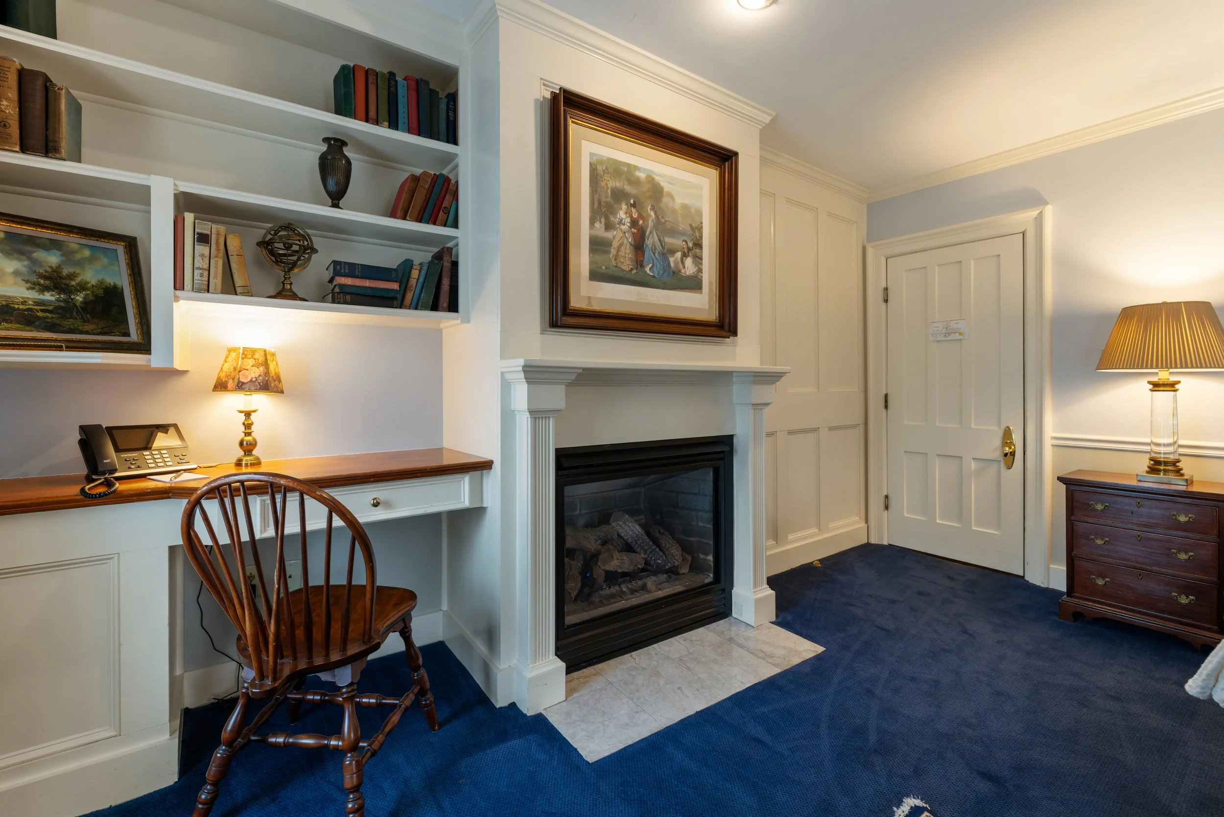 Interior of a room featuring a wooden desk with a telephone, a wooden chair, a white built-in bookshelf with books and decorative items, a fireplace with a framed painting above it, a brass desk lamp, and a table lamp with a pleated shade. The room has white walls, a blue carpet, and a white door.