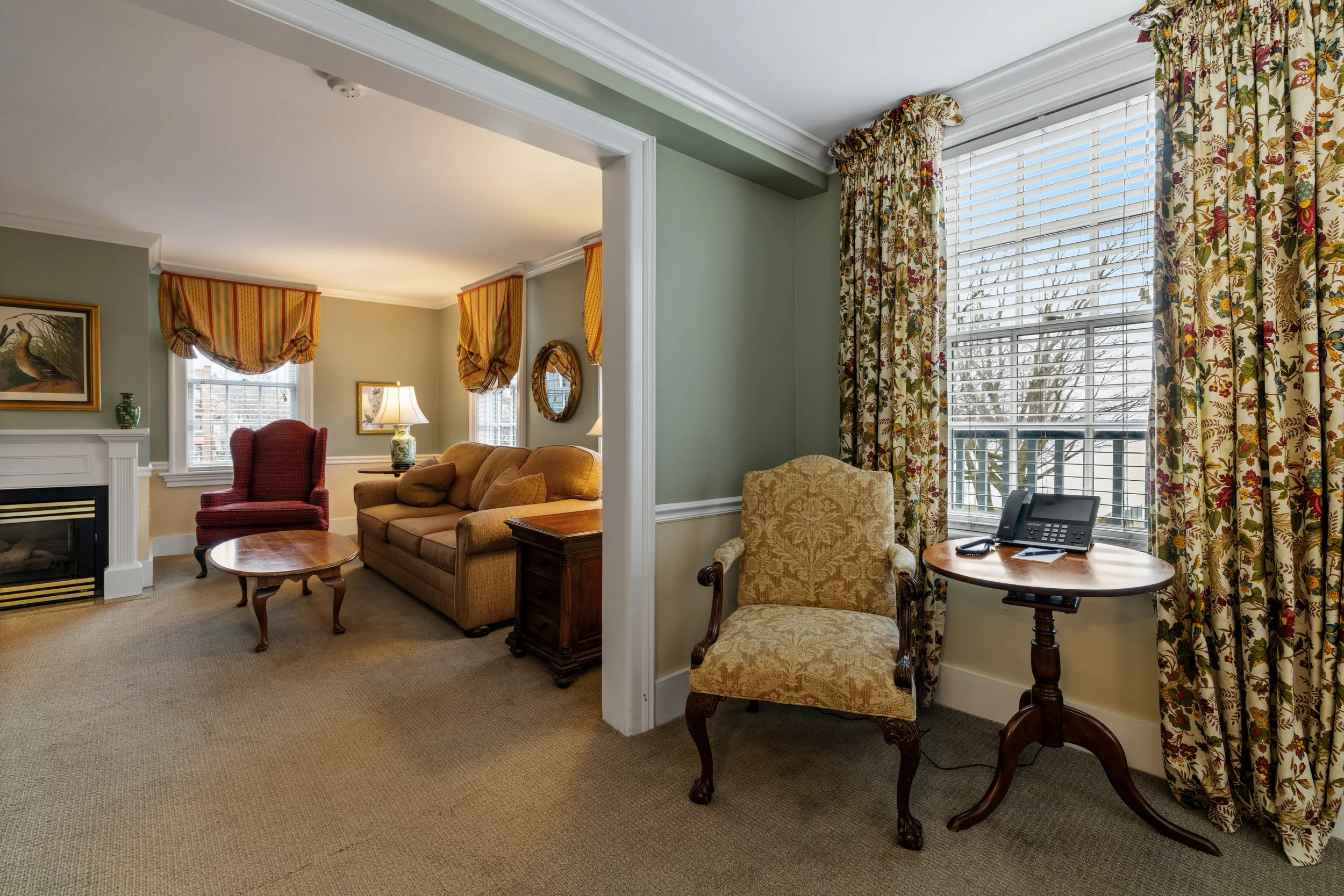 Living room with green walls, large windows with floral curtains, and traditional furniture including a sofa, armchair, and side tables.
