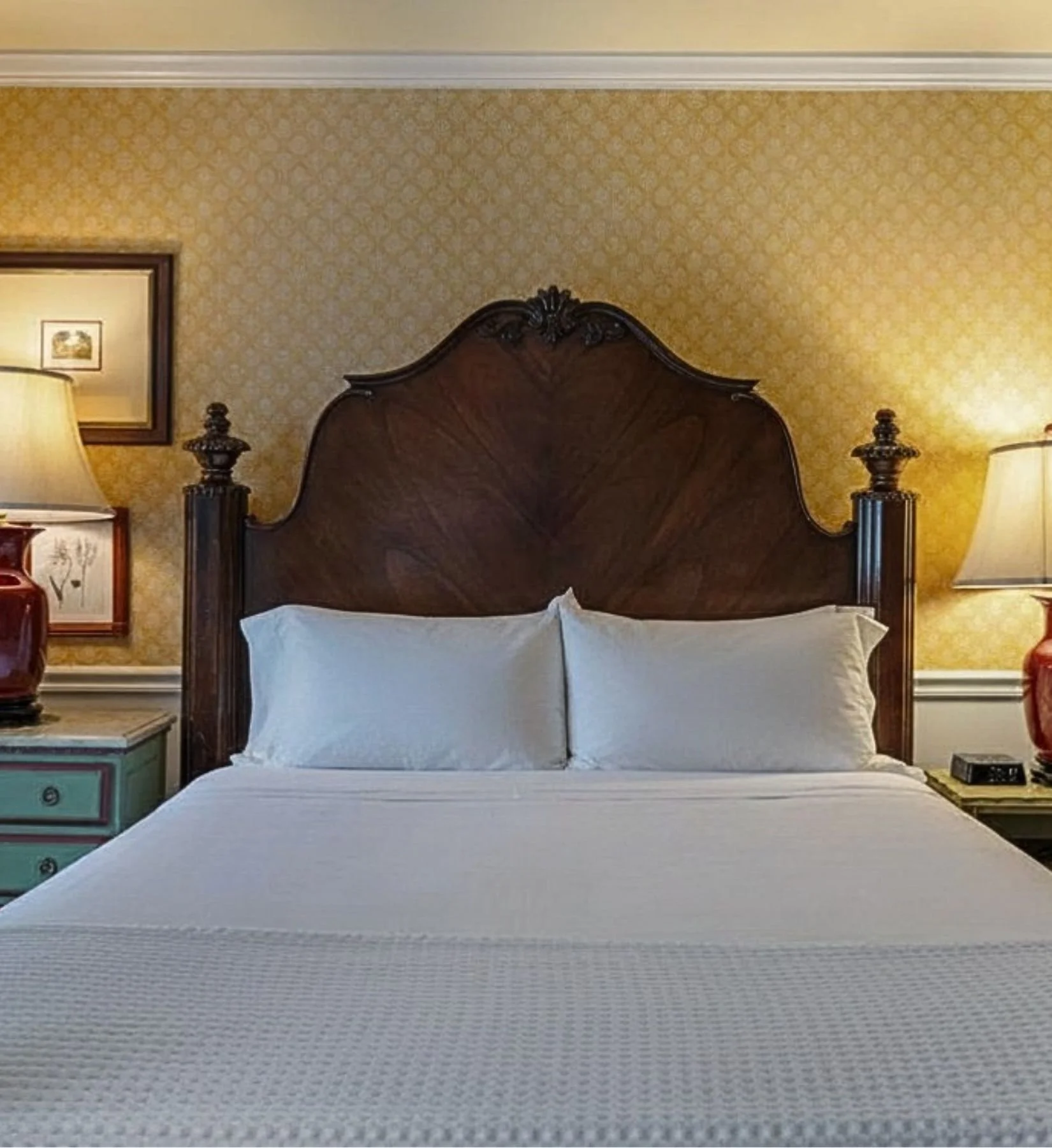 A neatly made bed with white sheets and pillows, a dark wooden headboard with ornate details, flanked by two matching nightstands with lamps, a framed art piece on the wall, and yellow patterned wallpaper in the background.