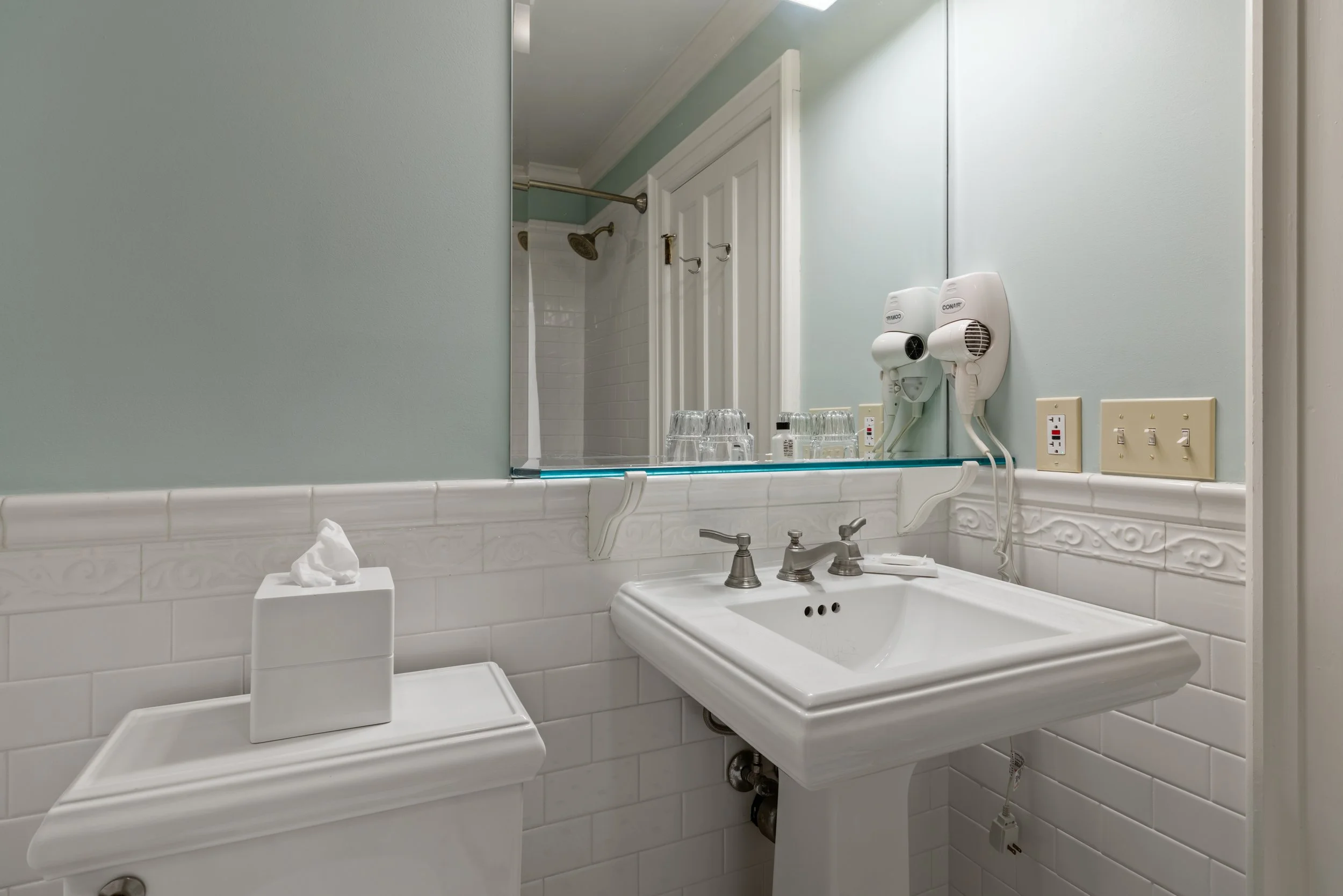 Bathroom mirror reflecting a bathroom with a shower, white sink, hairdryer, glasses, and electrical outlets.