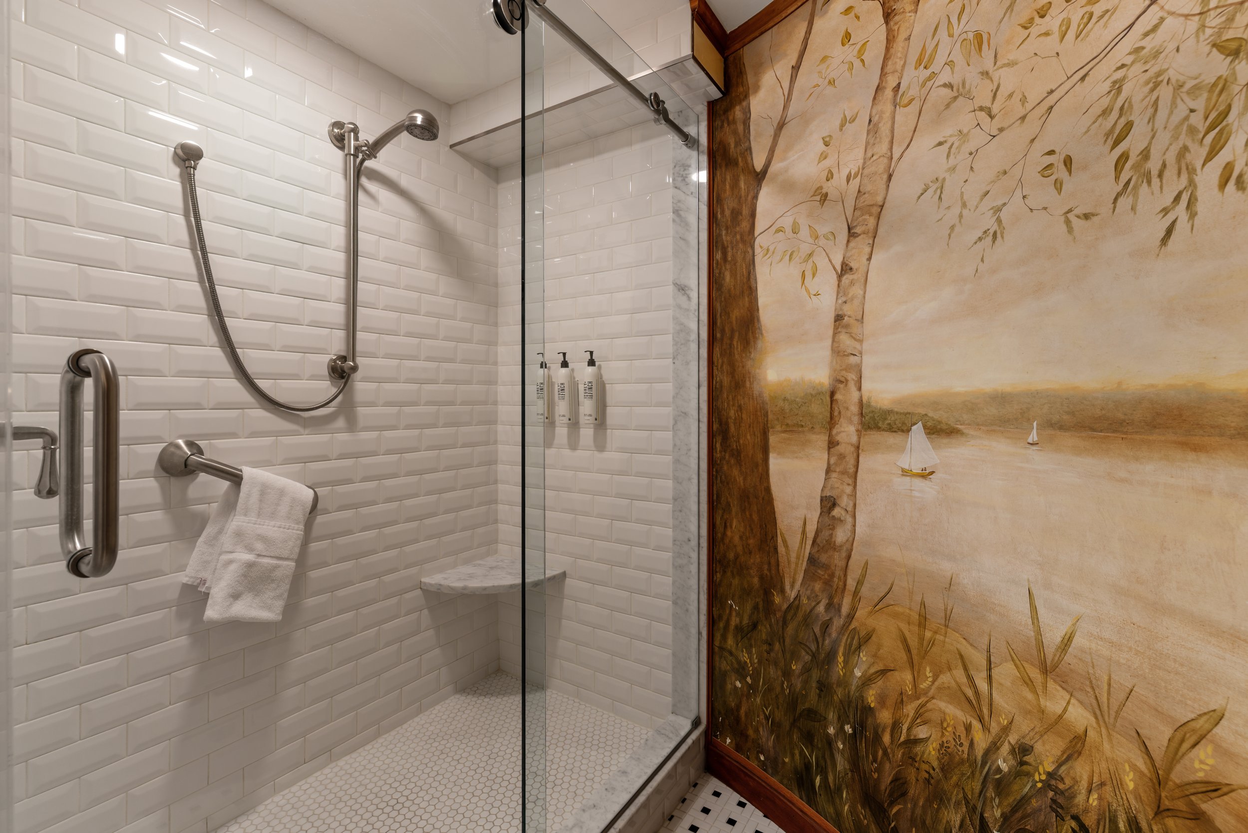 A bathroom shower area with white subway tiles, a handheld showerhead, a glass door, and a mural of trees and sailboats on the adjacent wall.
