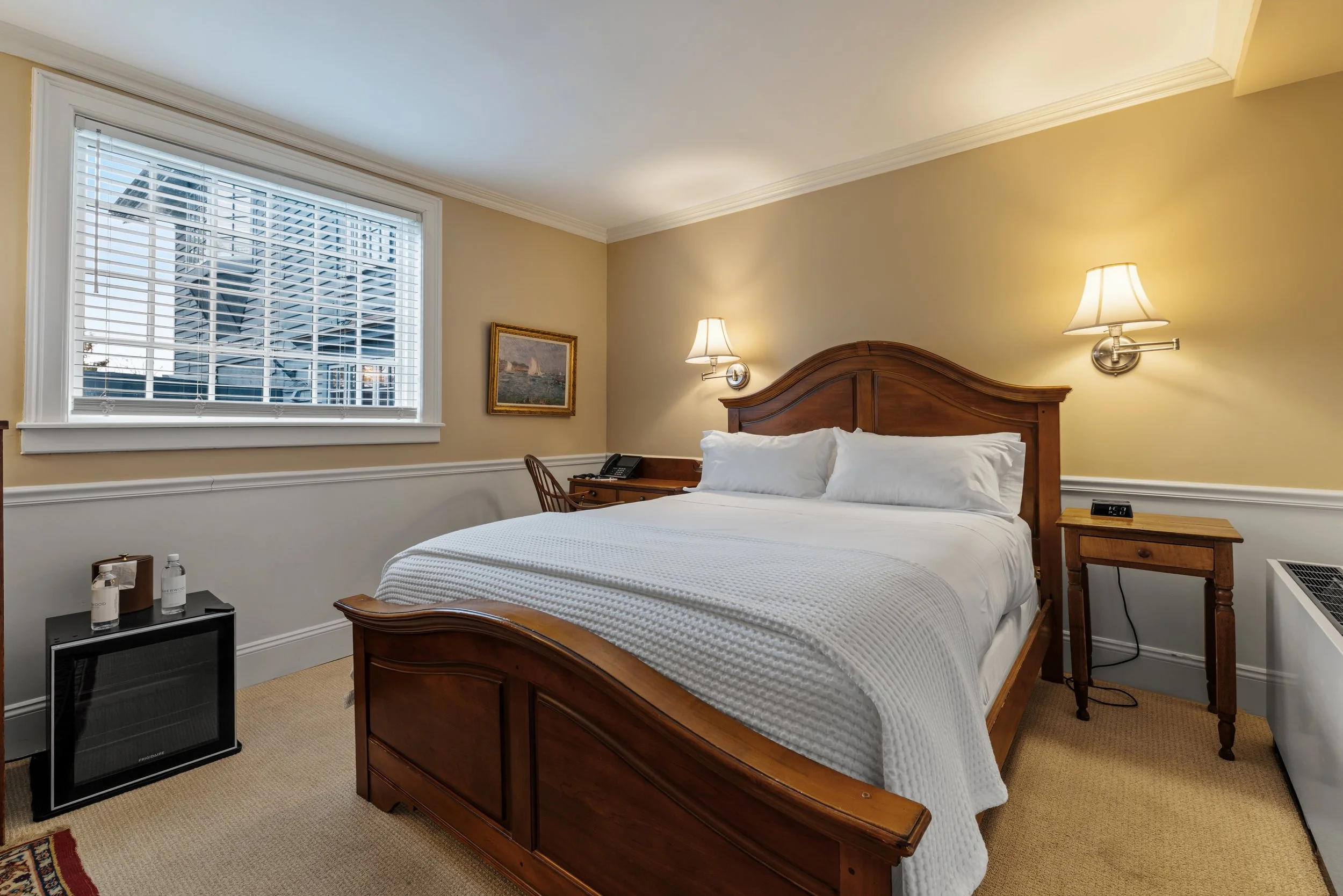 A hotel room featuring a wooden bed frame with white bedding, illuminated by two wall-mounted lamps, with a window showing a city view, a small side table with bottles and a mini fridge, and a framed painting on the wall.