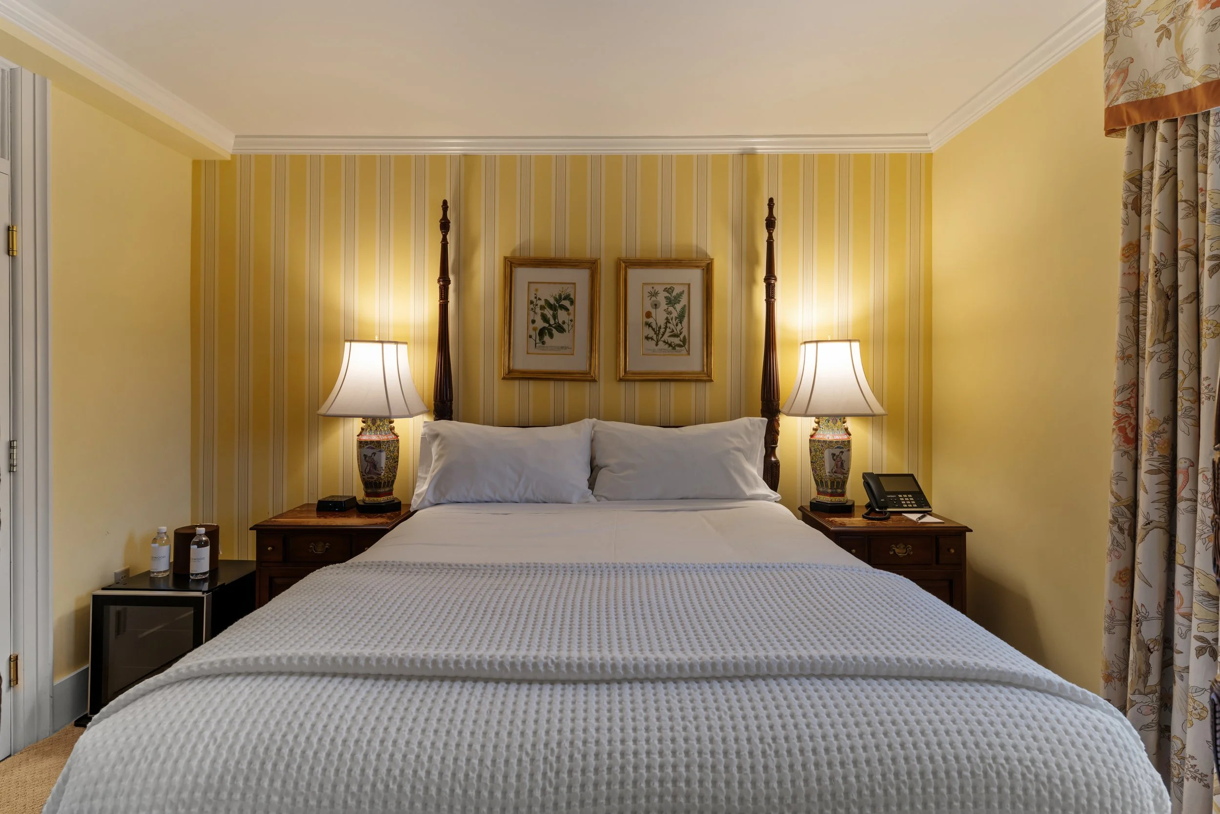 A hotel room with a bed, pillows, bedside tables with lamps, and decorative framed botanical prints on the yellow-striped wallpaper wall.