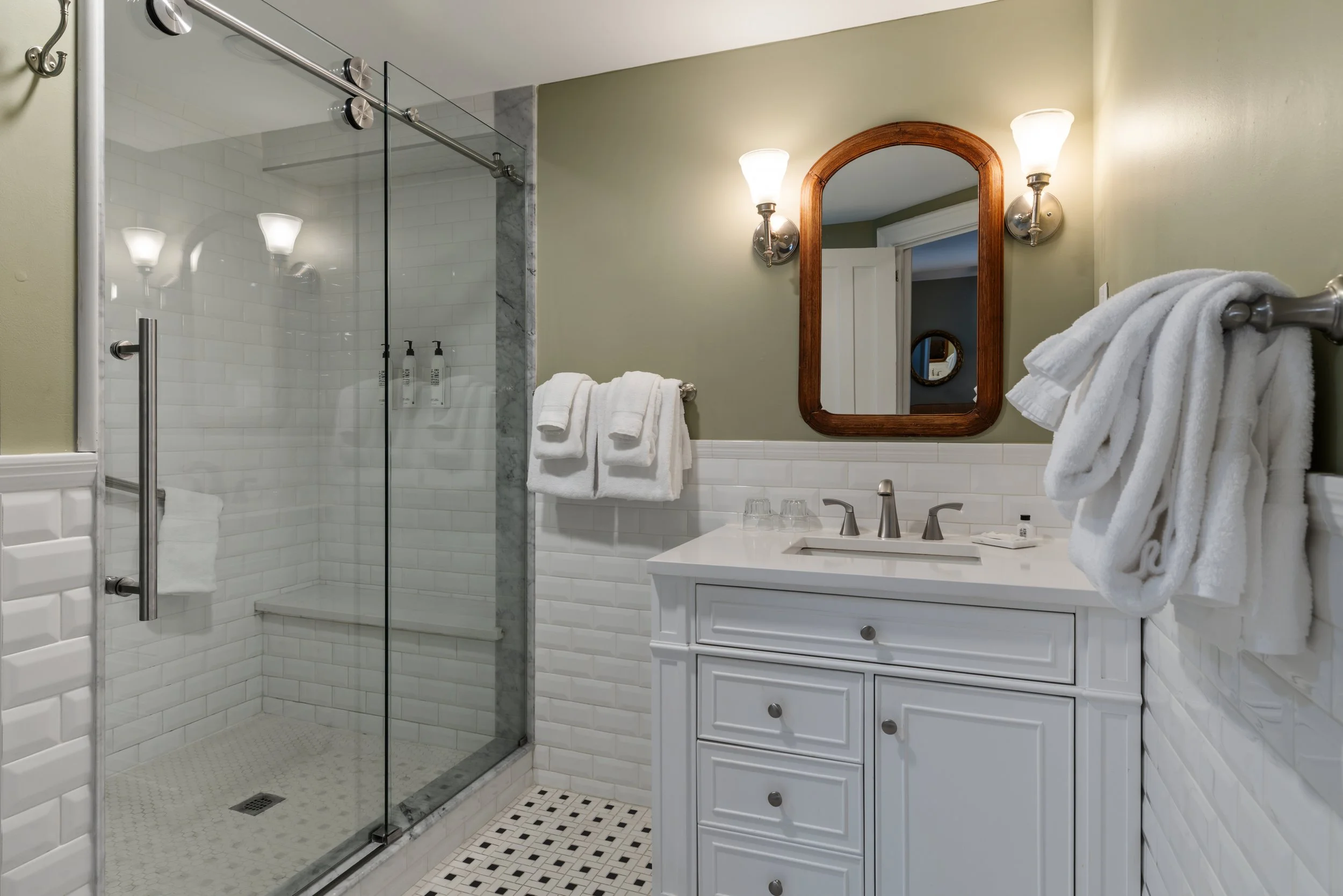 A bathroom with a shower area enclosed by glass, a white vanity with a sink, a mirror with a wooden frame, and two wall-mounted light fixtures. There are two white towels on a towel rack and a small tray with toiletries.