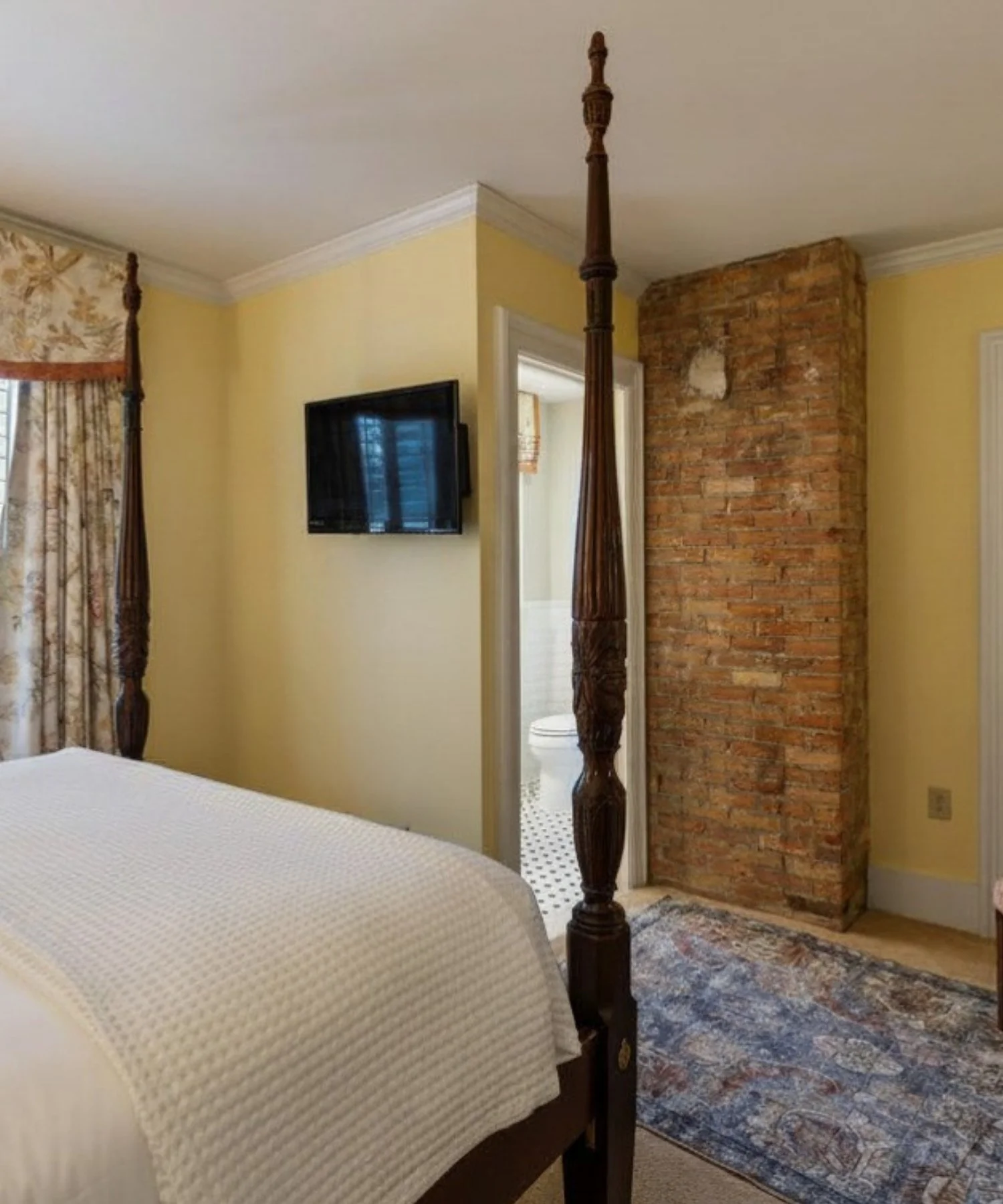 A bedroom with a four-poster bed, a yellow wall, a brick wall, a small rug, a wall-mounted TV, a floral curtain, and a doorway to a bathroom with a white toilet.