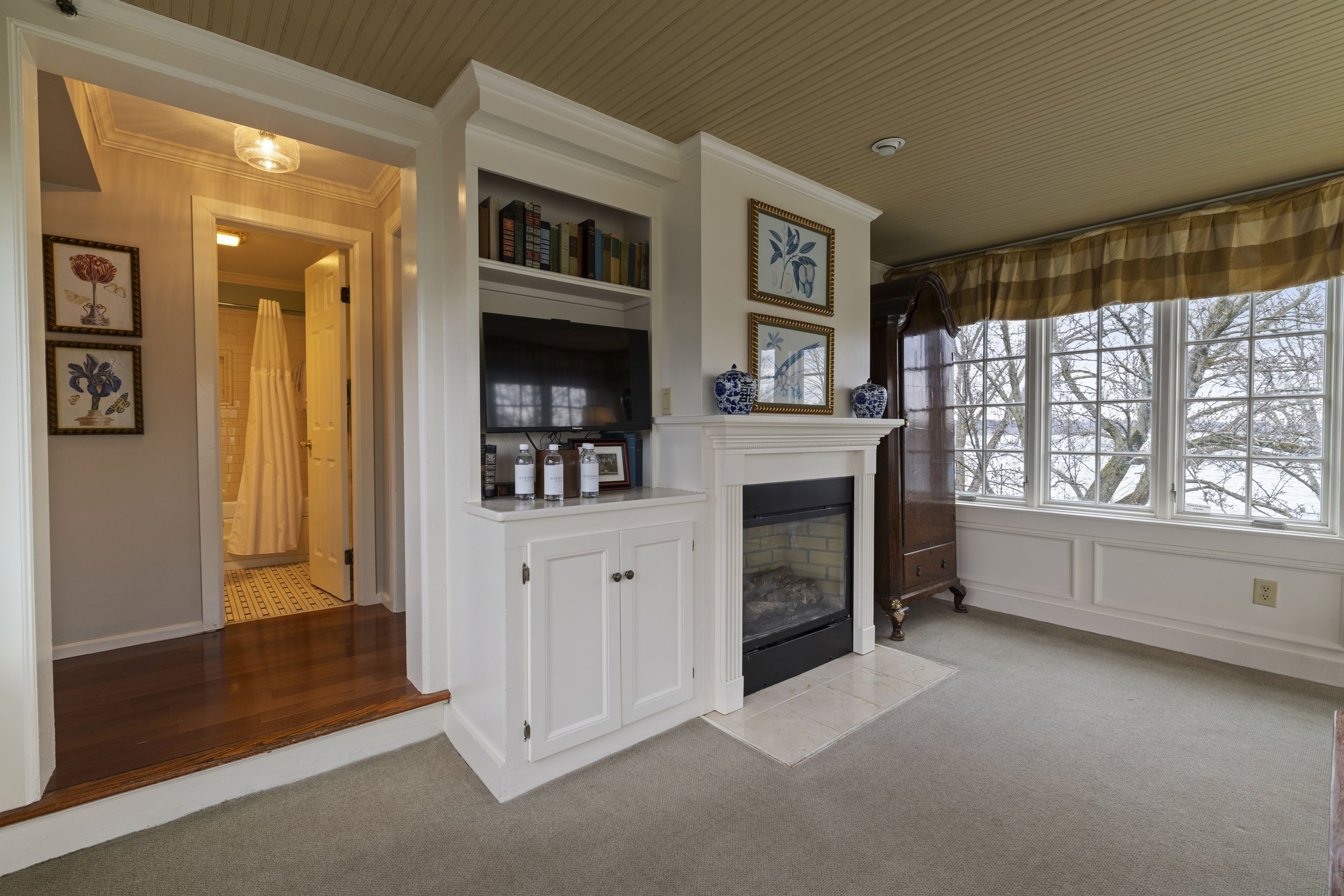 Living room with a fireplace, built-in bookshelf, framed art, and a large window with a yellow valance, showing trees outside.