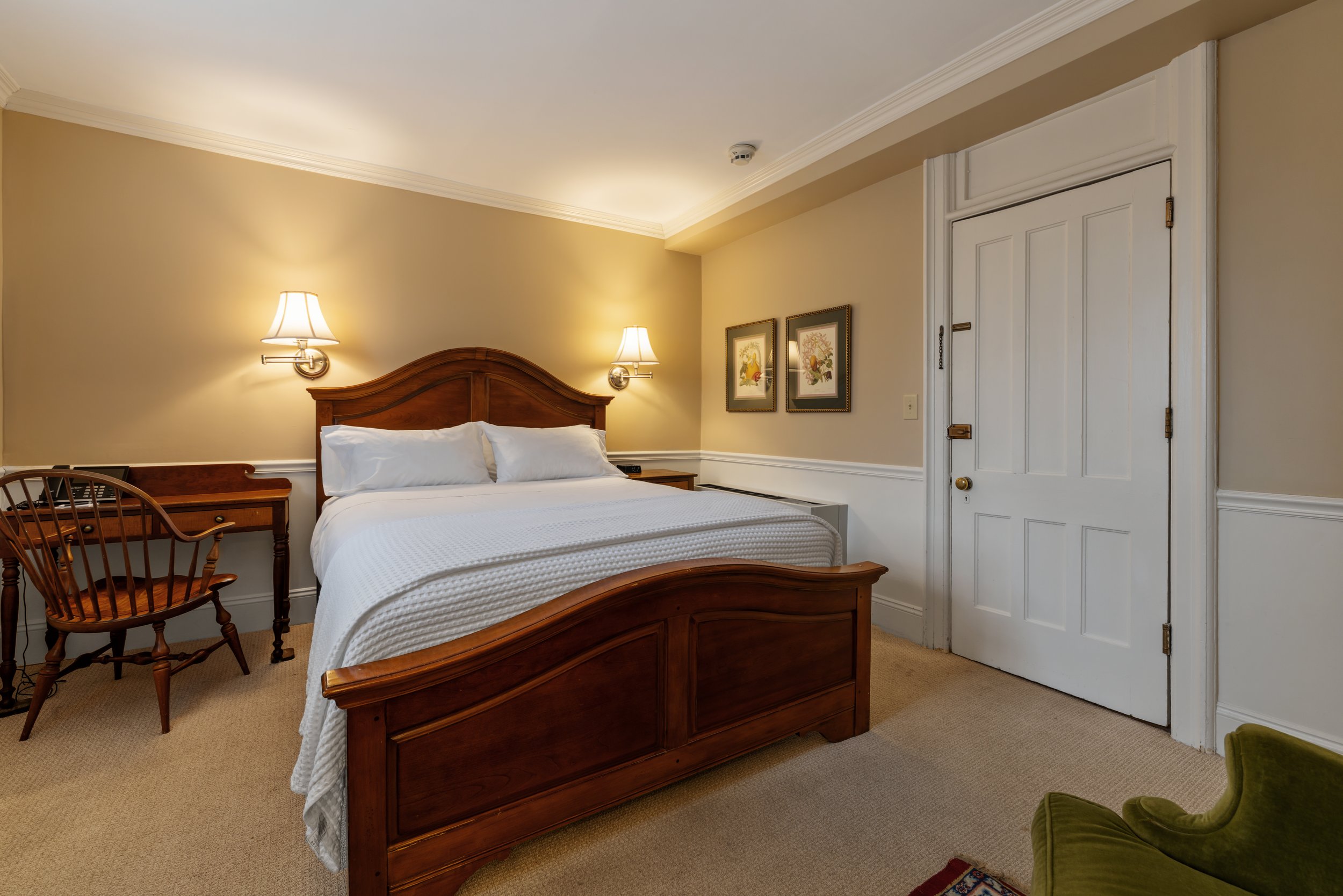 A hotel room with a wooden bed frame, two white bedsheets, and pillows. There are two wall-mounted lamps above the headboard. The walls are painted beige with decorative moldings at the ceiling. Two framed pictures of birds hang on the wall beside a closed white door with a brass doorknob and chain lock. A wooden chair with armrests is beside the bed, and a green armchair is partially visible in the bottom right corner.