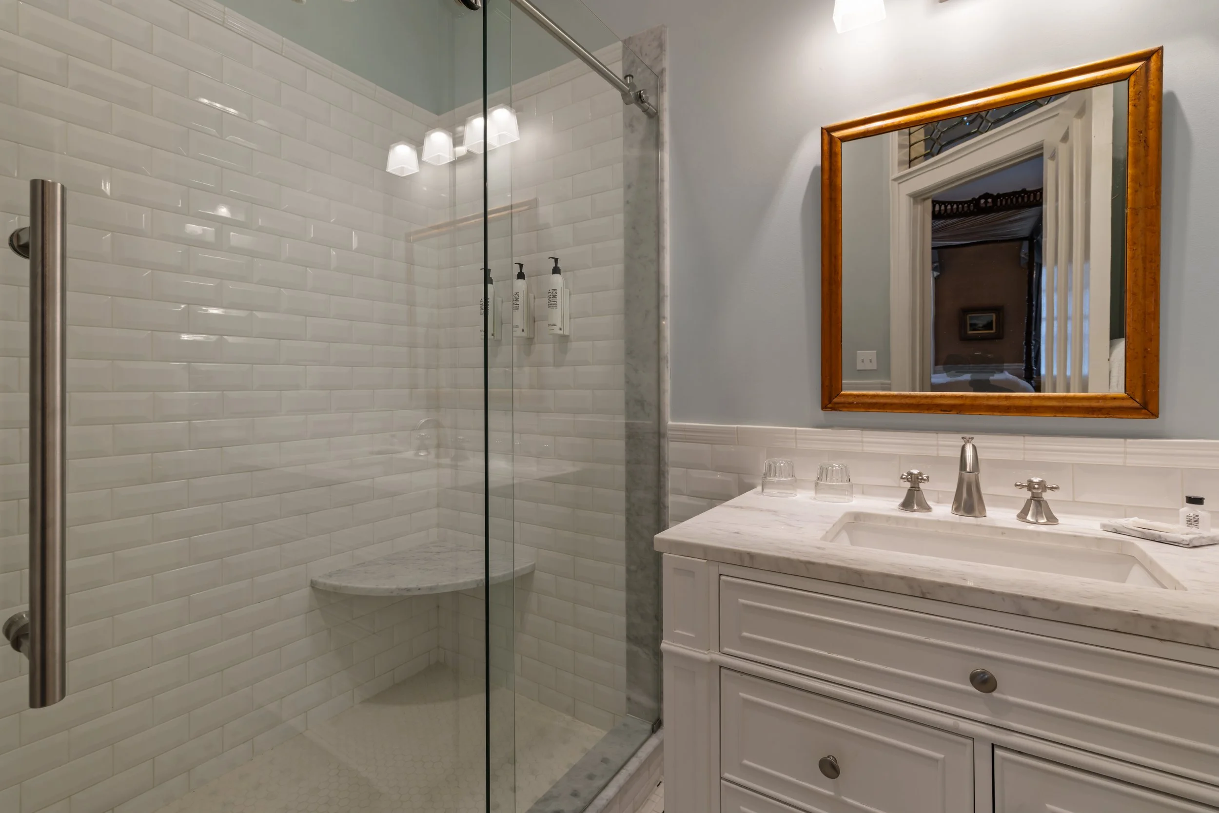 Bathroom with a walk-in shower featuring white subway tiles, a marble bench, and a glass door. There is a white vanity with a marble countertop, a mirror with a wooden frame, and wall-mounted light fixtures.