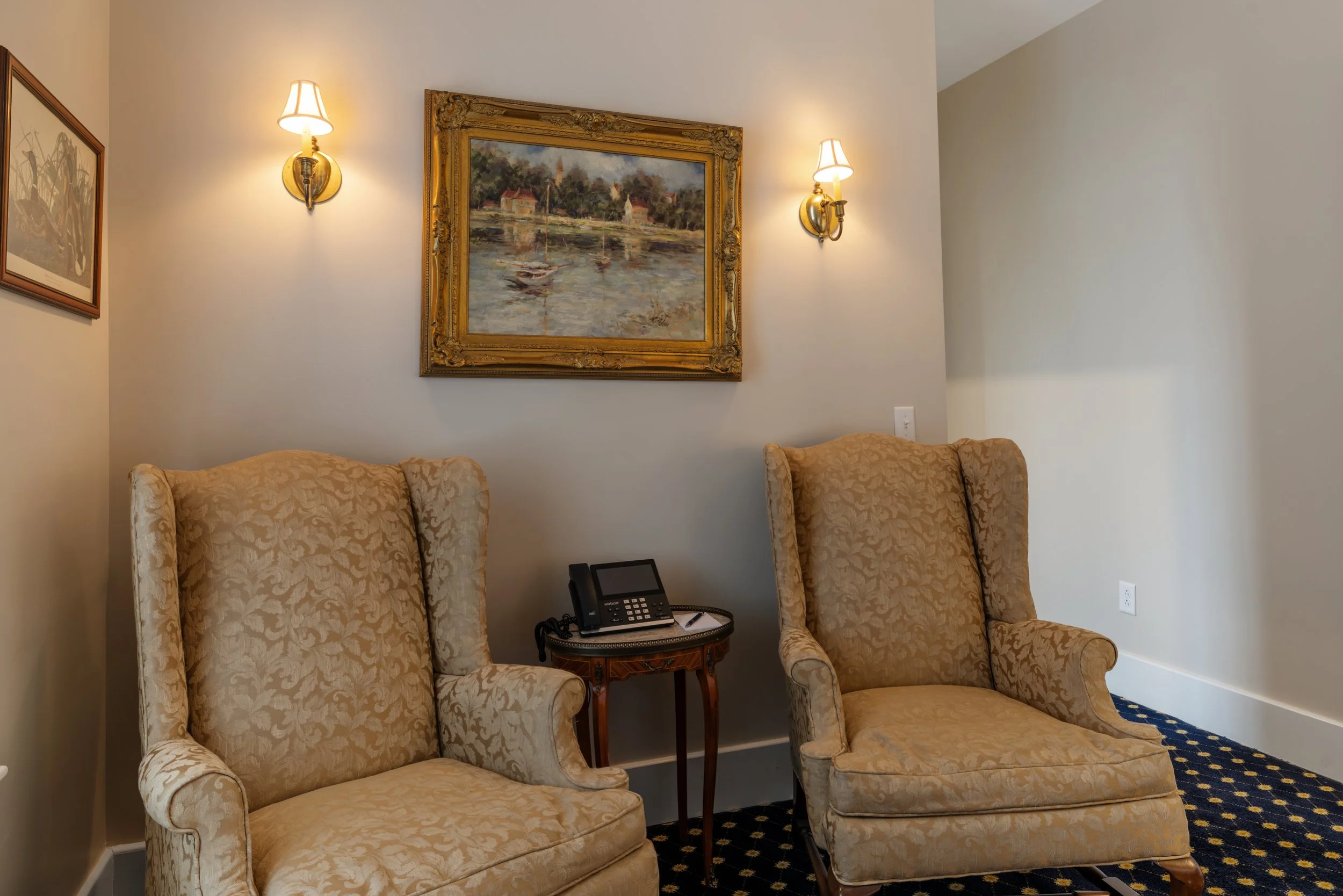 Two upholstered armchairs with floral fabric and matching footstool, a small round wooden side table with a black telephone and notepad, framed artwork on the wall, and wall-mounted lamps in a cozy room.