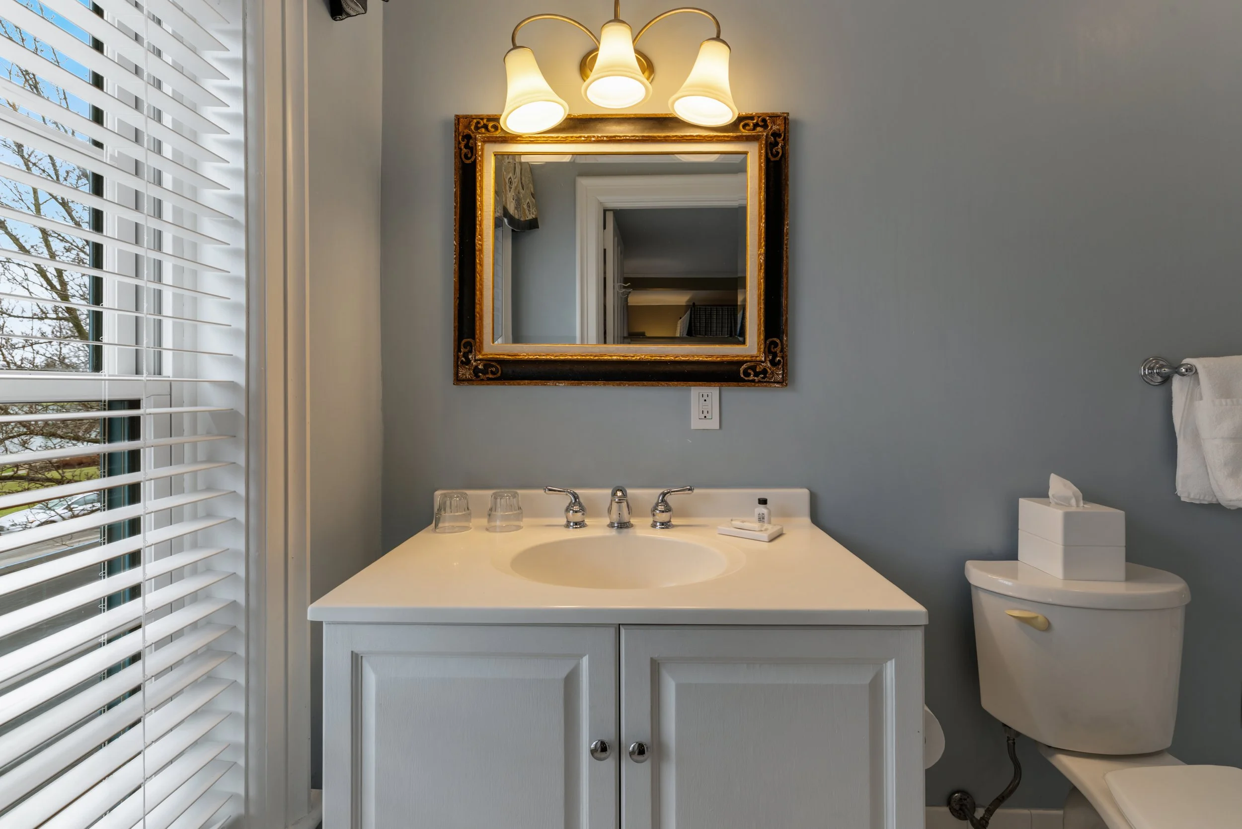 A bathroom with a window and white blinds on the left, a vanity with a sink, faucet, and mirror in the center, and a toilet on the right. The walls are painted light gray, and there is a decorative framed mirror above the sink. The bathroom has a light fixture with three lamps above the mirror.