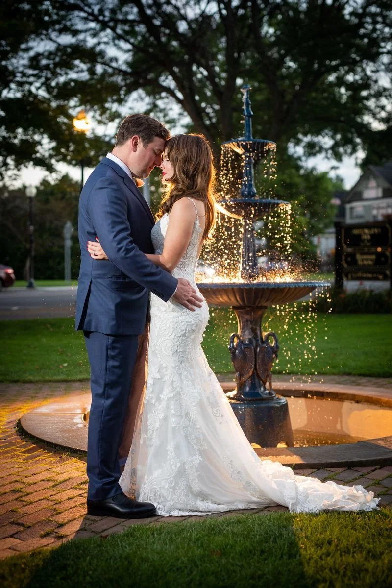 A bride and groom stand close together with foreheads touching by a fountain at sunset, surrounded by greenery and trees.