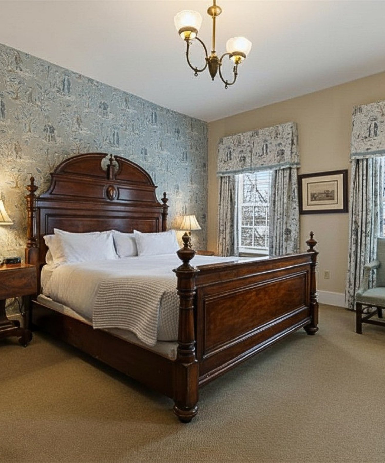 A bedroom with a large wooden bed, white bedding, and twin bedside lamps. The walls have a stone pattern and curtains with a matching pattern, and there is art on the wall between the windows. A ceiling light fixture hangs from the ceiling.