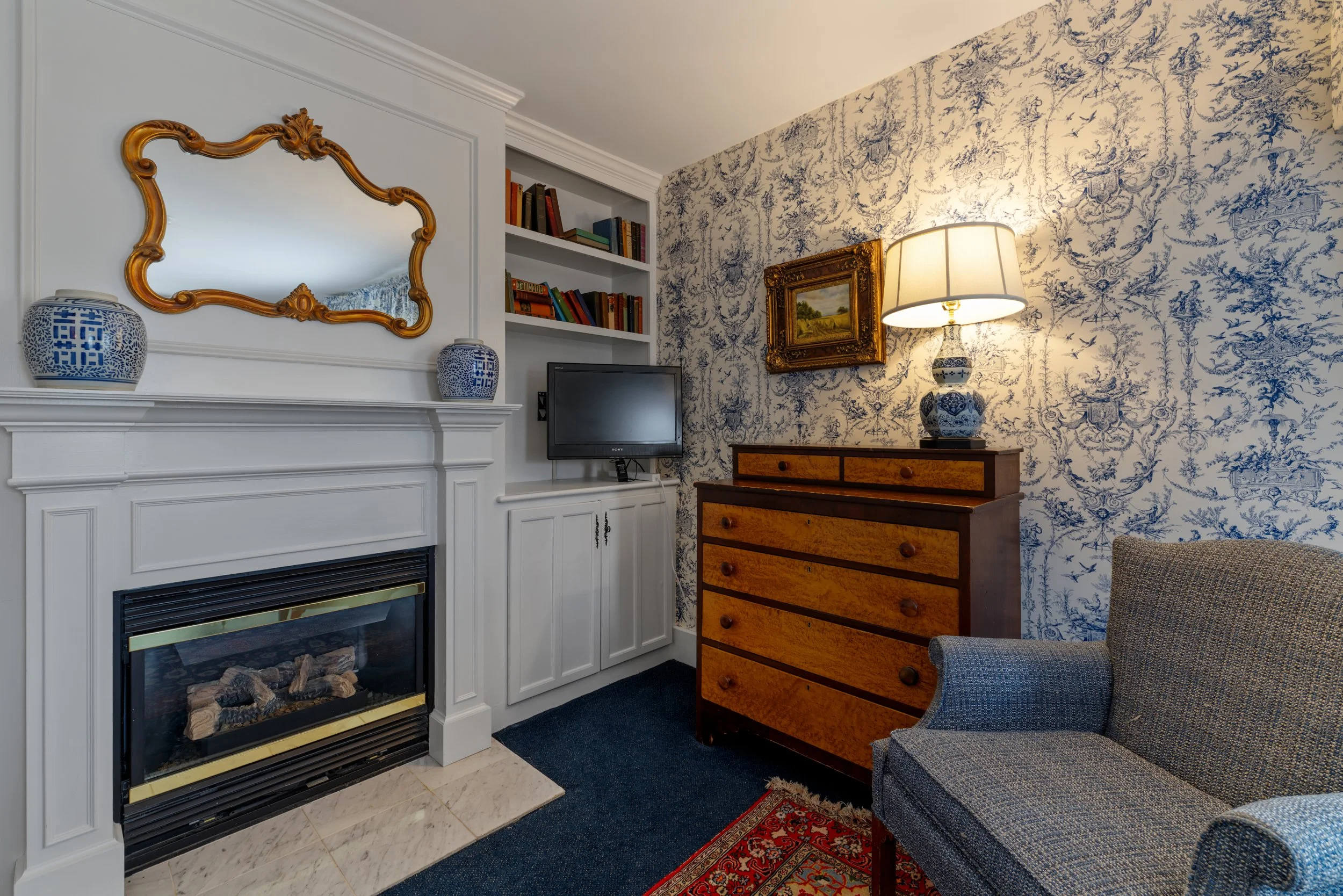 Living room with blue and white wallpaper, a white fireplace with a gold mirror above it, two blue and white vases on the mantle, a small flat-screen TV, a wooden chest of drawers, a table lamp, a painting in a gold frame, a gray armchair, and a red patterned rug.