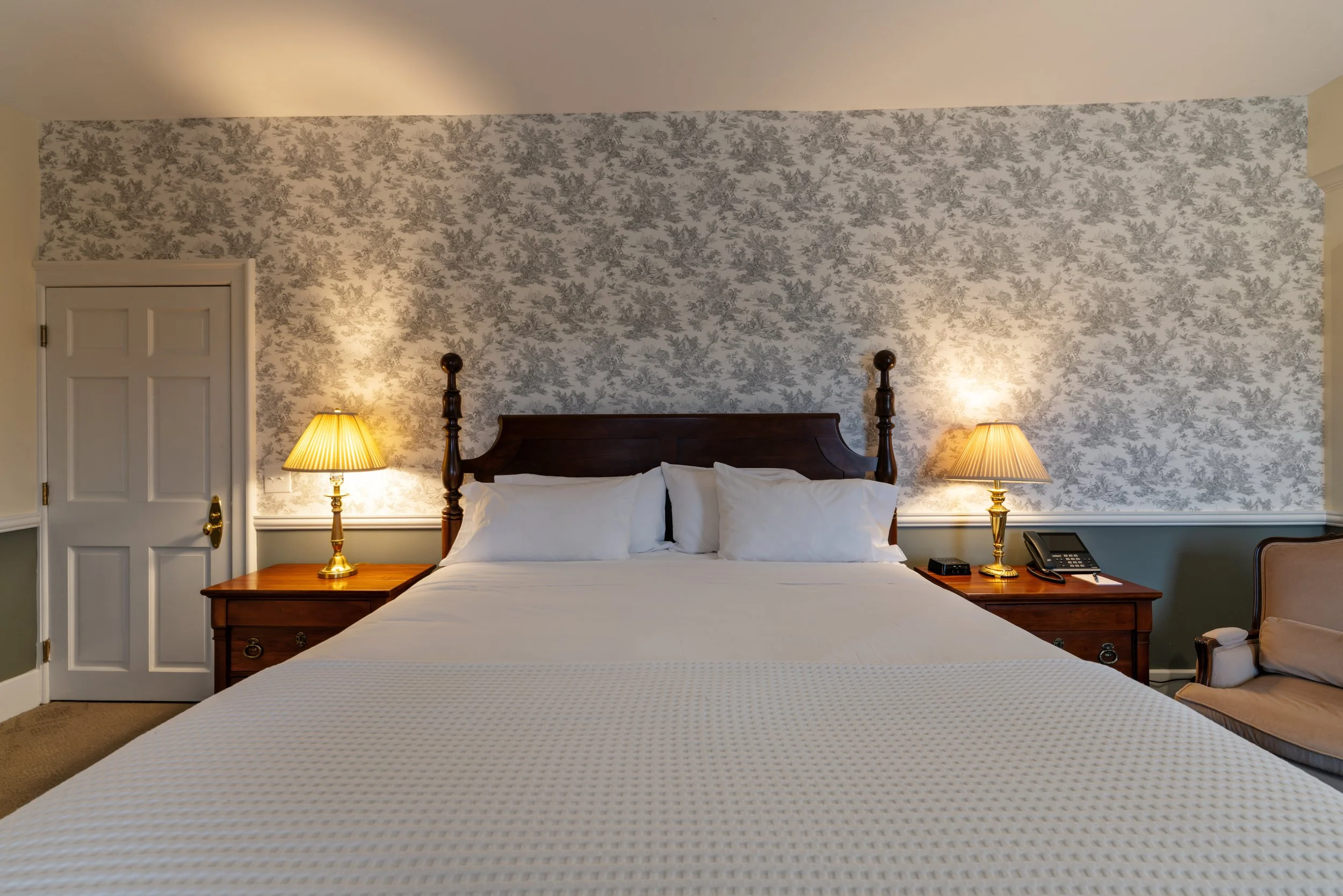 Hotel room with a large bed, wooden headboard, two bedside tables with lamps, and a wallpapered accent wall behind the bed.